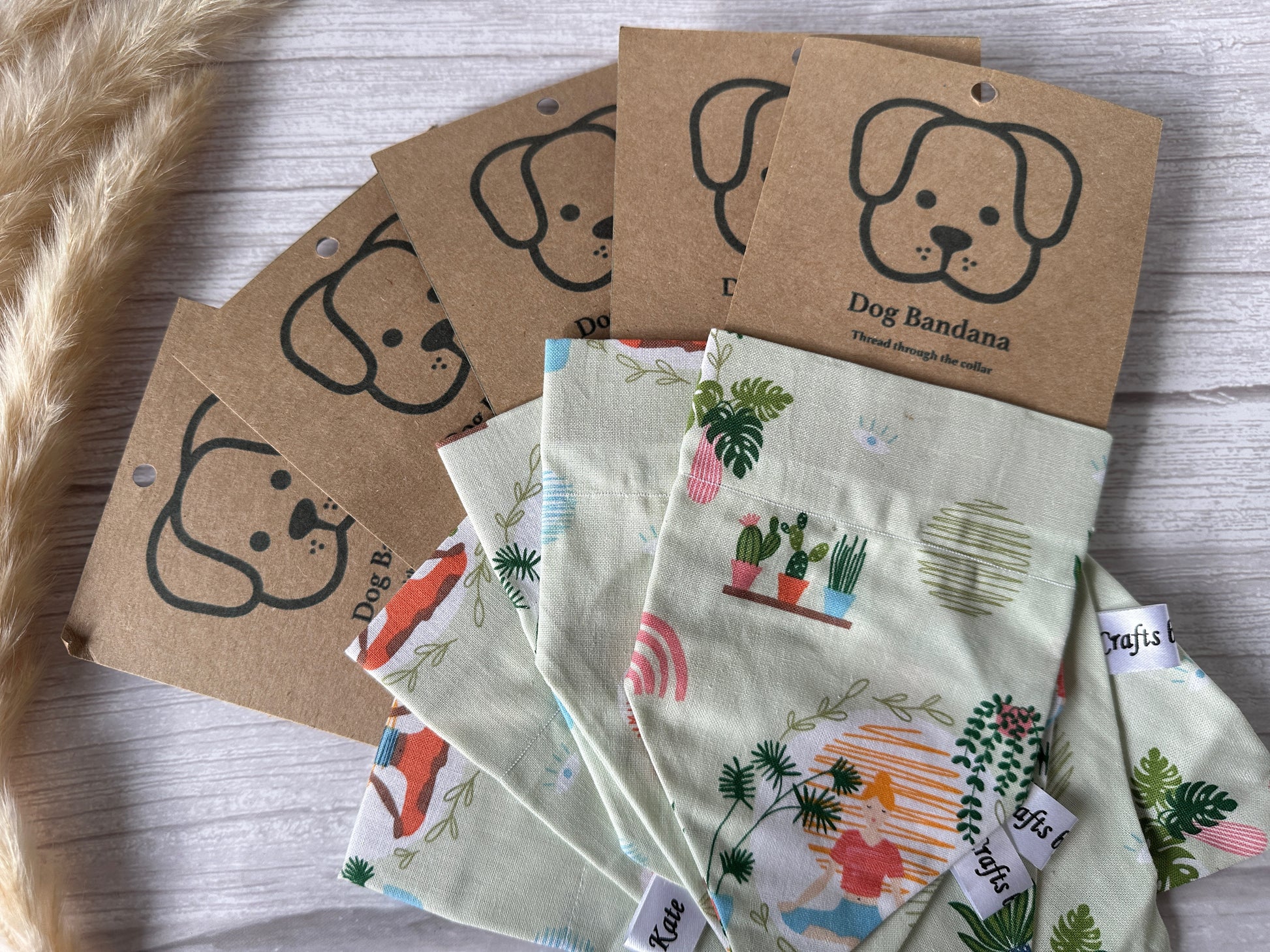 A collection of Cotton Pet Bandanas with collar attachments from Crafts by Kate lies arranged in a fanned-out pattern on a light wooden surface. Each bandana, designed for plant and yoga lovers, features various colorful patterns and comes with kraft paper packaging displaying a cute dog illustration and the text "Dog Bandana." Dried wheat stalks are to the left.