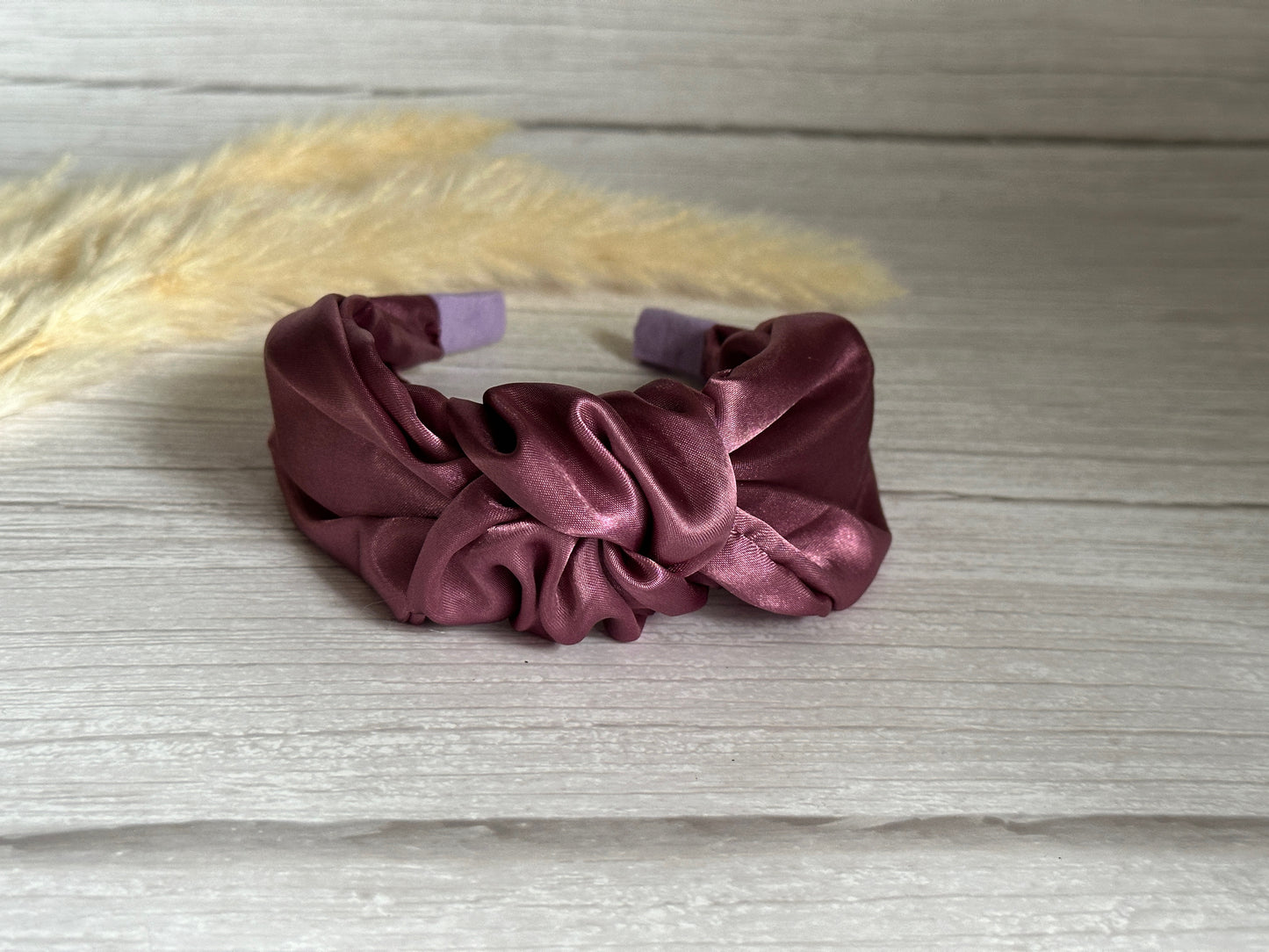 A luxurious, handcrafted Silk Knotted Headband in plum purple by Crafts by Kate is showcased against a light wooden background, accented with dried grass in the corner.