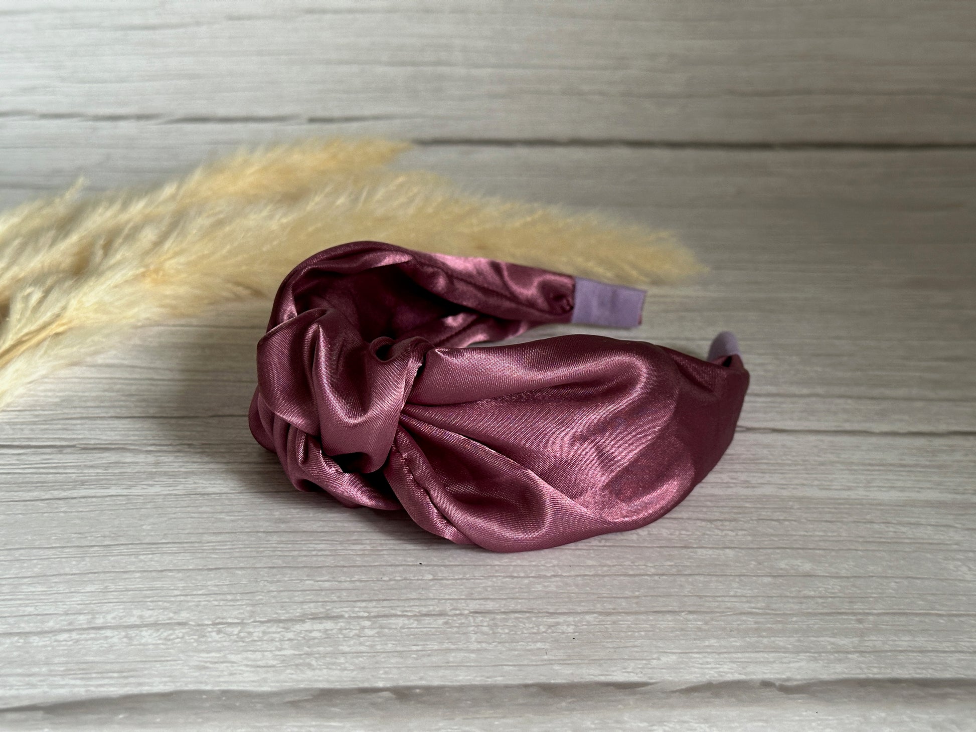 A luxurious touch from Crafts by Kate, the Silk Knotted Headband in a rich plum purple color elegantly rests on a light wooden surface. Soft, beige pampas grass stems in the background add an air of sophistication.