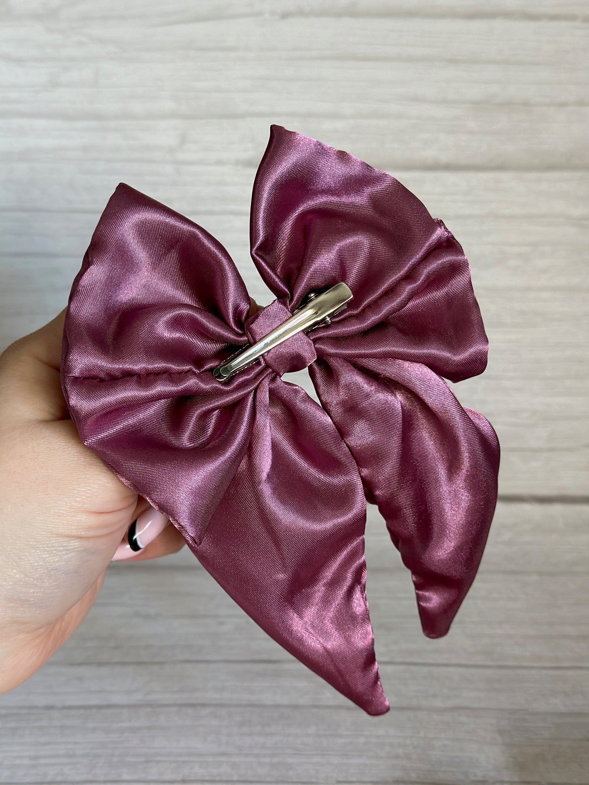 A hand holding the luxurious Silk Sailor Bow Clip in a rich plum purple shade from Crafts by Kate, featuring a sleek silver clip, is set against an elegant light wood-textured background.