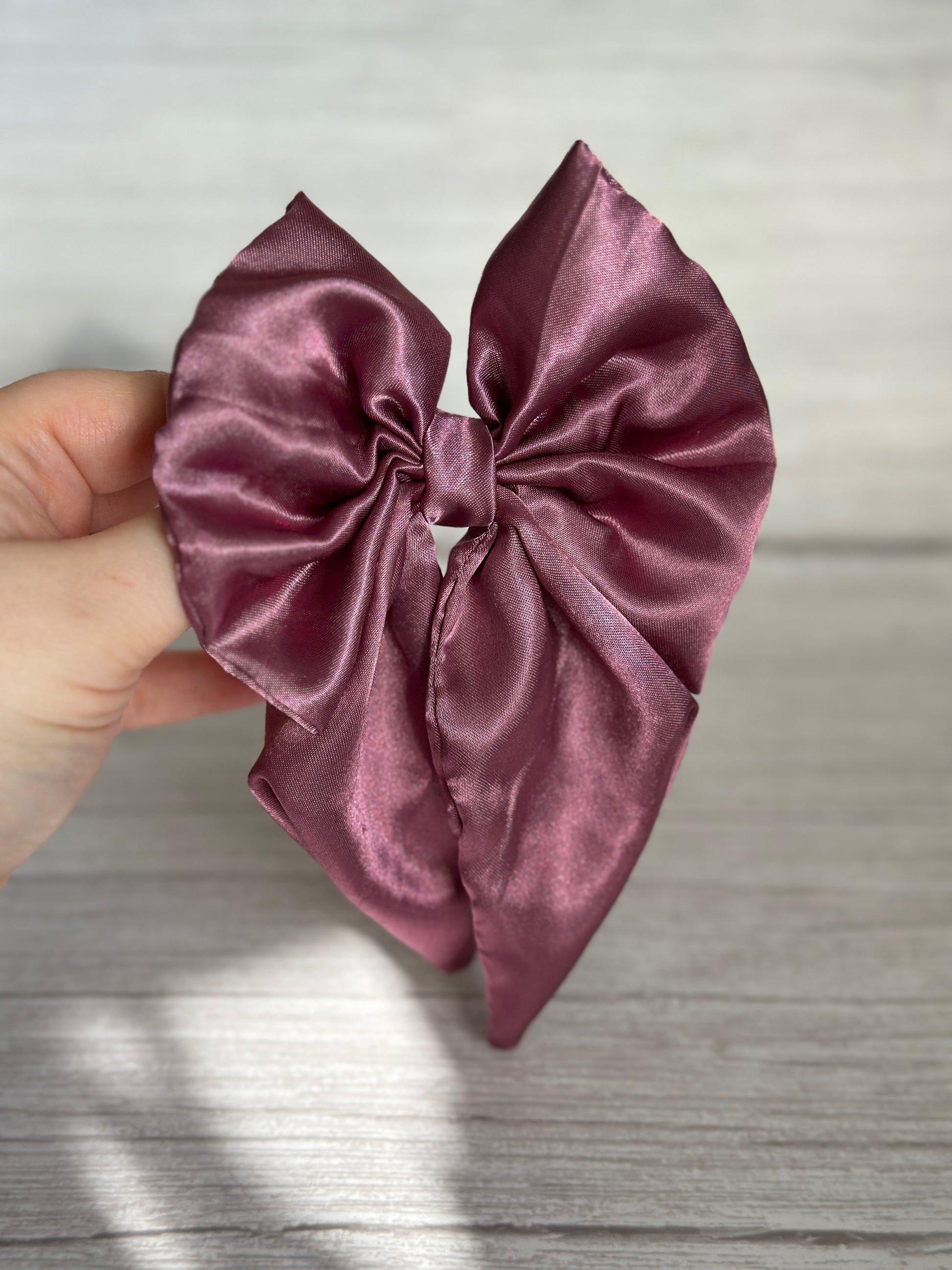 A hand holds a luxurious, glossy clip from Crafts by Kate—a Silk Sailor Bow in a rich plum purple shade—against a light wooden background.