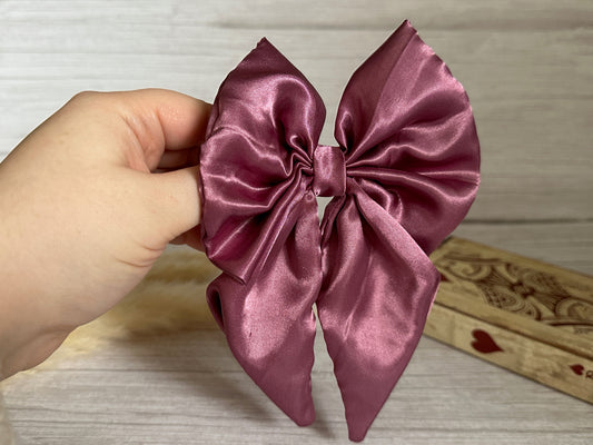 A hand elegantly holds the luxurious Silk Sailor Bow Clip in a rich plum purple from Crafts by Kate, its silky folds and tails gleaming. The background is a neutral, textured surface, with a wooden item featuring a heart design partially visible on one side.