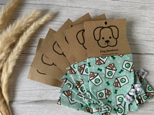 A stack of green handcrafted "Cotton Pet Bandana with collar attachment - Pupcups" from Crafts by Kate, featuring a sushi pattern and labeled "Dog Bandana," with brown tags that have illustrations of a dog's face. These bandanas are displayed on a light-colored wooden surface, accented by some wheat stalks to the side.