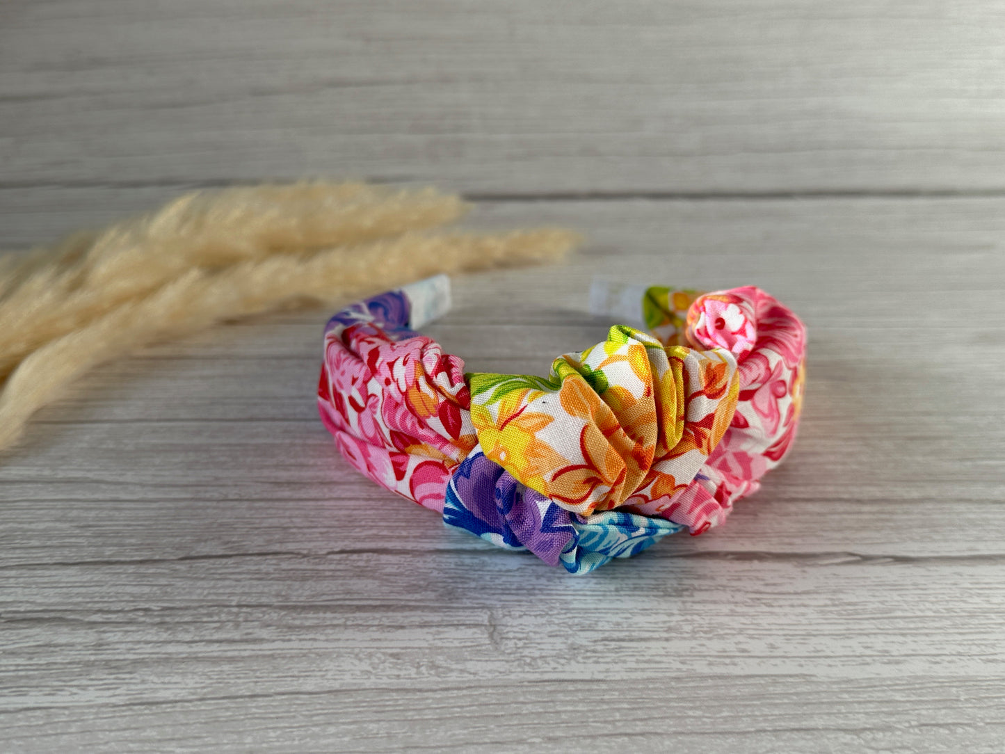 A vibrant Cotton Knotted Headband - Rainbow Floral by Crafts by Kate, adorned with a large bow and featuring vivid colors including pink, yellow, and blue, is placed on a light wood surface. Adjacent to it, soft beige pampas grass adds a touch of natural contrast.