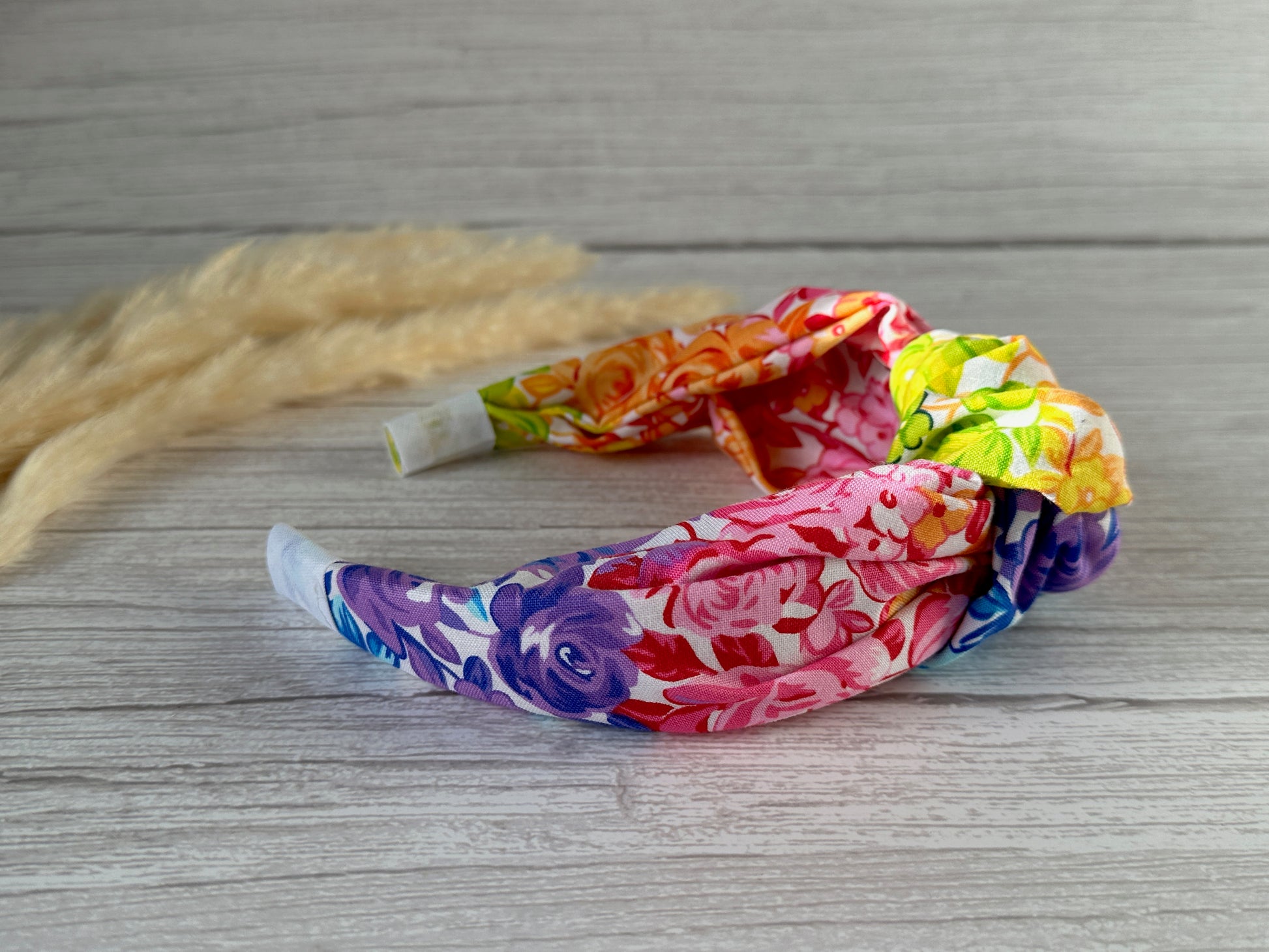 A vibrant, floral-patterned Cotton Knotted Headband - Rainbow Floral from Crafts by Kate lies coiled on a light wooden surface. This headband showcases lively shades of purple, pink, orange, and green with delicate flowers printed across the fabric. In the background are light beige dried decorative grasses.