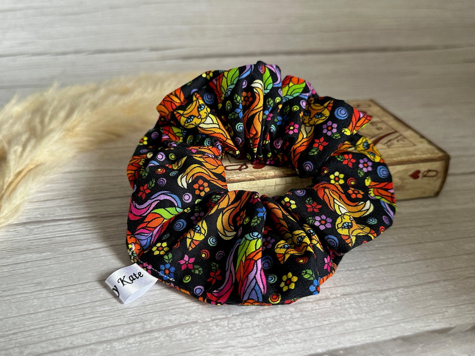 An eye-catching, handcrafted luxury scrunchie featuring the vibrant Rainbow Ginger Cat design set against a black background is displayed on a wooden surface. A small white tag with the name "Crafts by Kate" is attached to the extra-large Cotton Scrunchie. A light-colored, feathery decor piece is partially visible in the background.