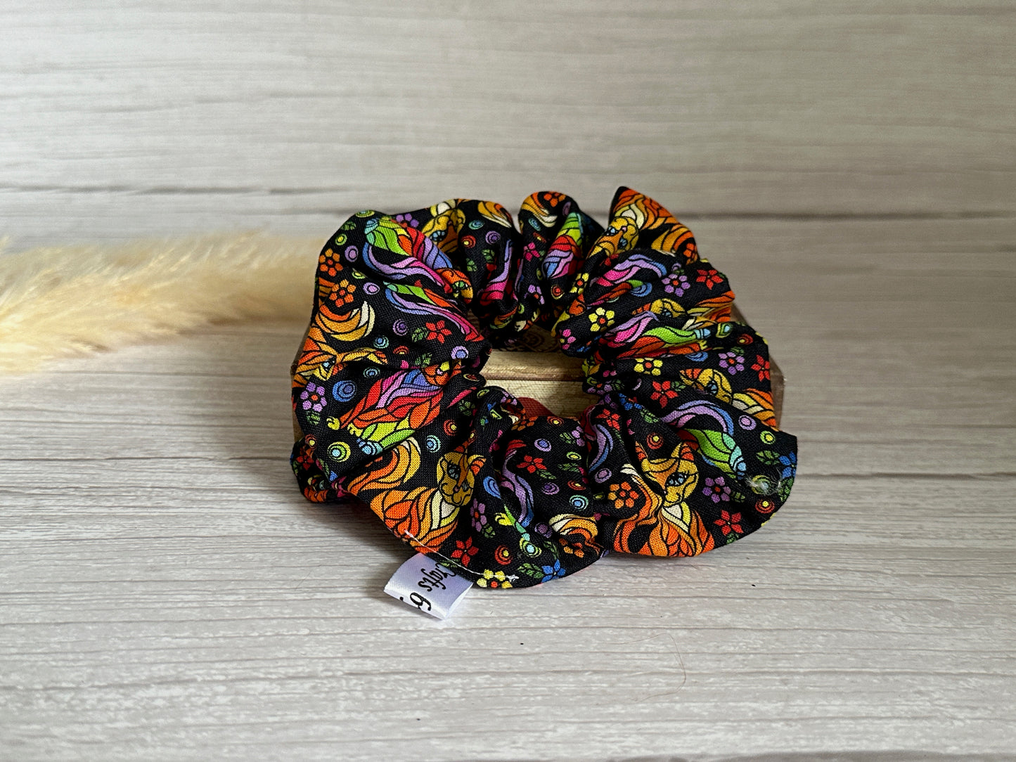 The Cotton Extra Large Scrunchie - Rainbow Ginger Cat by Crafts by Kate is a handcrafted luxury accessory featuring a vibrant pattern with multicolored swirls, leaves, and flowers. Displayed on a light-colored wooden surface, it offers a clear view of its intricate design. This colorful scrunchie also features a small white tag.