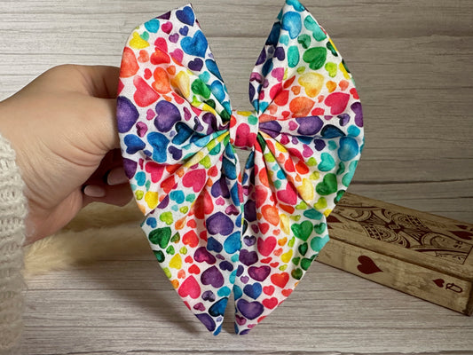 A hand is holding the Cotton Sailor Bow Clip - Rainbow Hearts, a large handcrafted luxury bow by Crafts by Kate. The bow features a vibrant display of hearts in various shades of rainbow colors on high-quality cotton fabric. It is set against a light wood background, and a wooden box with playing card designs can be seen in the background.