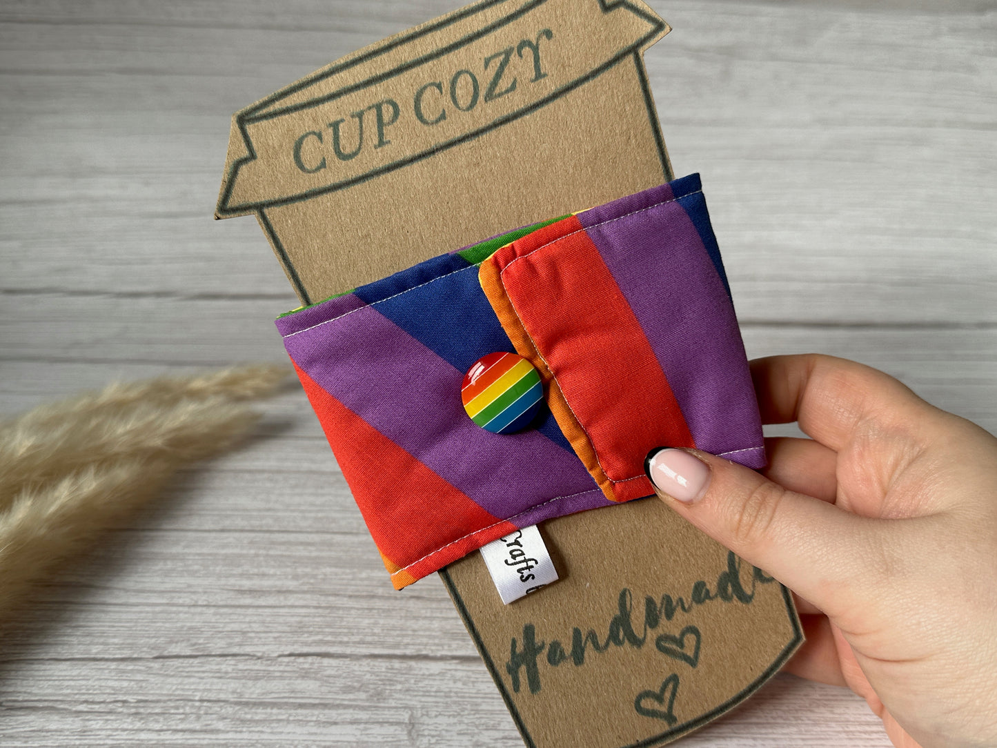 A person is holding a vibrant, handcrafted Cotton Reusable Cup Cozy - Rainbow Stripes from Crafts by Kate. The cozy features a rainbow-colored button and is wrapped around a cardboard cutout shaped like a coffee cup with the words "CUP COZY" and "handmade" written on it. This eco-friendly coffee sleeve adds an environmentally conscious touch, with dried grass in the background.