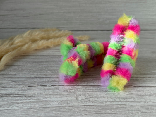 Two vibrant, fluffy caterpillar-shaped accessories from Crafts by Kate rest on a light wooden surface. Branded as the Fluffy Hair Claw Clip - Rave Rainbow, these luxury plush fabric clips feature a mix of green, pink, yellow, and purple hues. To the left, there are some beige tufted fibers resembling natural dried grasses.