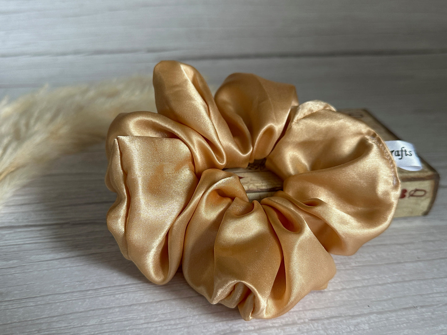 A Silk Extra Large Scrunchie in the Rich Gold shade from Crafts by Kate rests elegantly on a light wooden surface. In the background, a small wooden box and a piece of dried pampas grass enhance its luxurious charm.