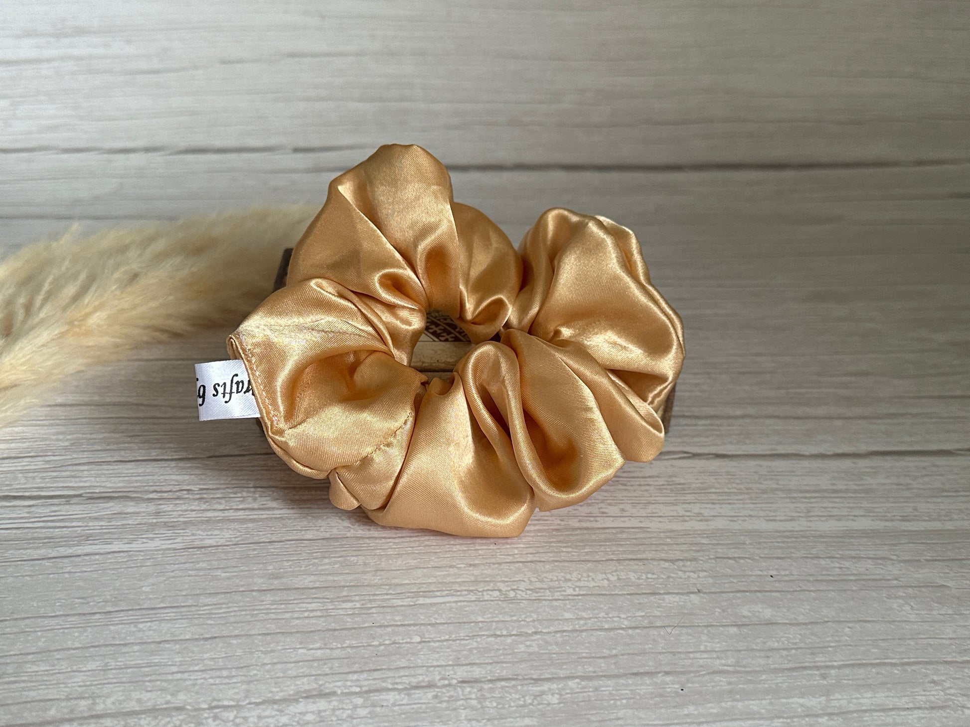 A luxurious Silk Extra Large Scrunchie in a rich golden hue from Crafts by Kate rests on a wooden surface, complete with a small white tag.