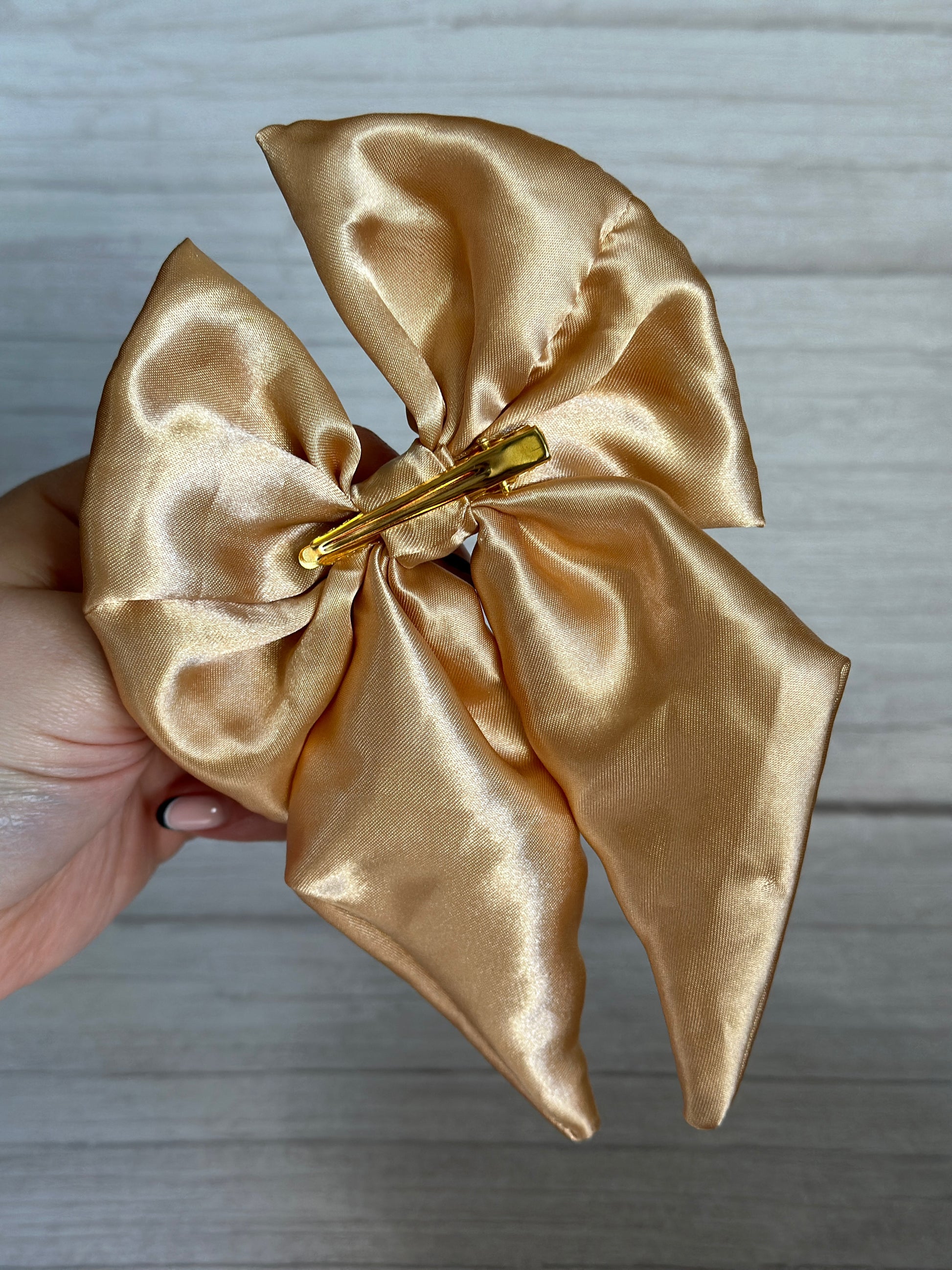 A hand presents a luxurious Silk Sailor Bow Clip in Rich Gold from Crafts by Kate against a light wood background. This carefully handcrafted bow showcases its smooth and elegant texture, featuring a large clip at the center.