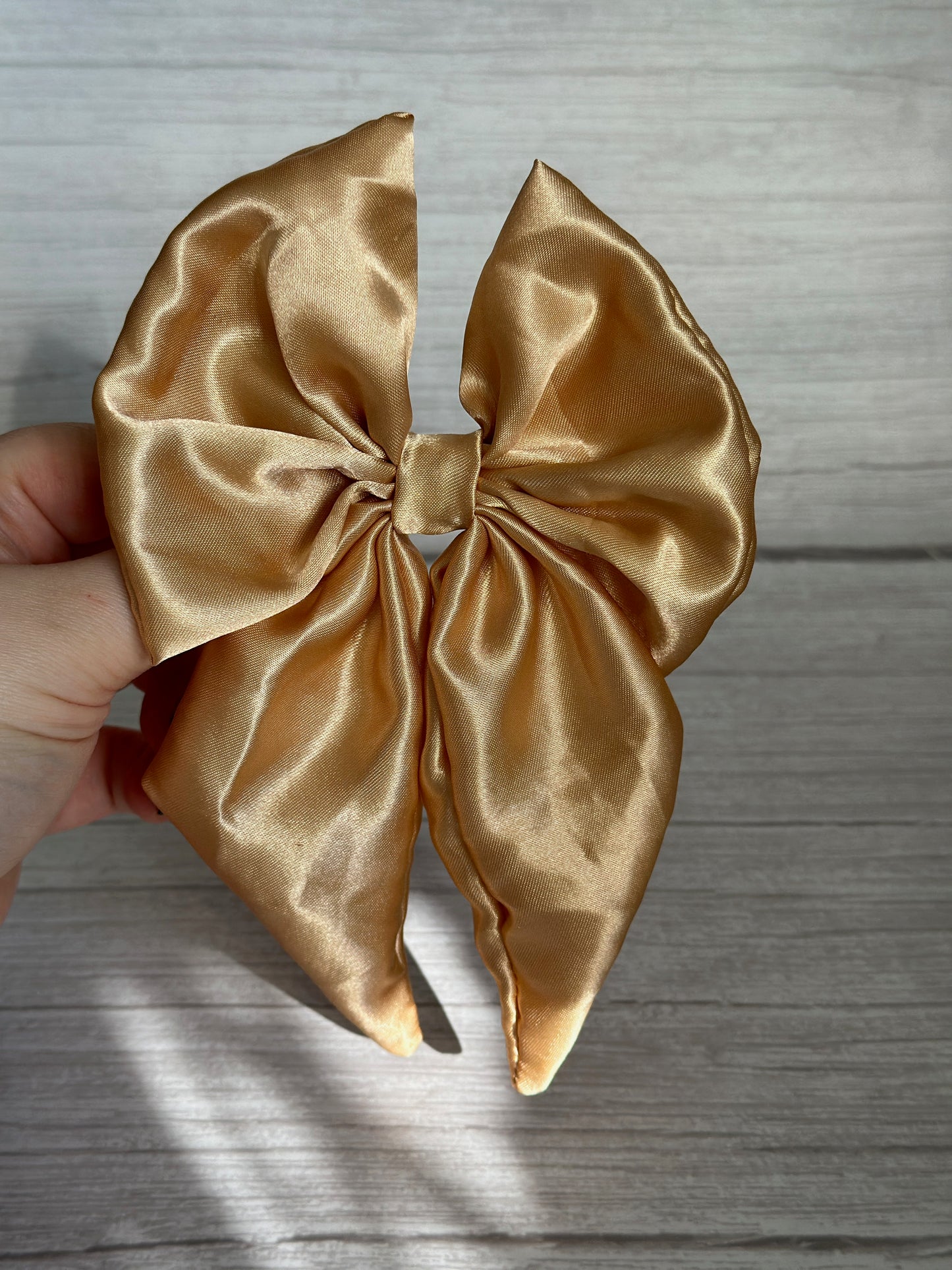 A hand holds the luxurious Silk Sailor Bow Clip - Rich Gold by Crafts by Kate against a wooden background. Its shiny texture and neatly tied loops and tails exude an elegant handcrafted charm.