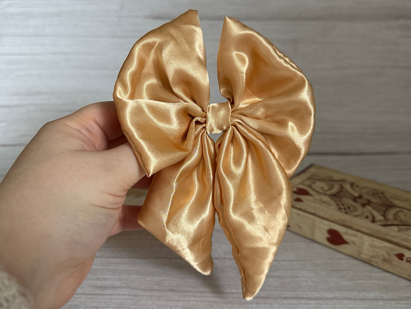 A hand artfully showcases the Silk Sailor Bow Clip in Rich Gold from Crafts by Kate, set against a wooden background. In the vicinity, a decorative box featuring heart designs adds charm, enhancing the elegance of this exquisite bow clip ensemble.