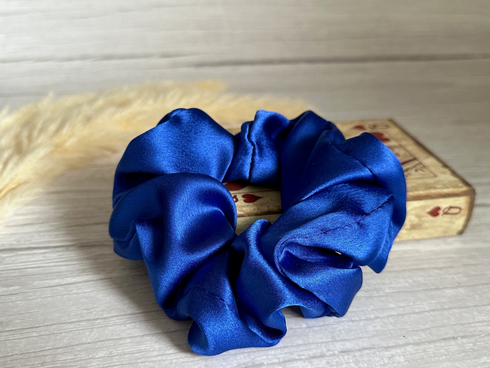 A Silk Extra Large Scrunchie in royal blue from Crafts by Kate rests gracefully atop a deck of cards, highlighted by its ace design. In the softly blurred background, beige pampas grass adorns the light wooden surface with an understated elegance.