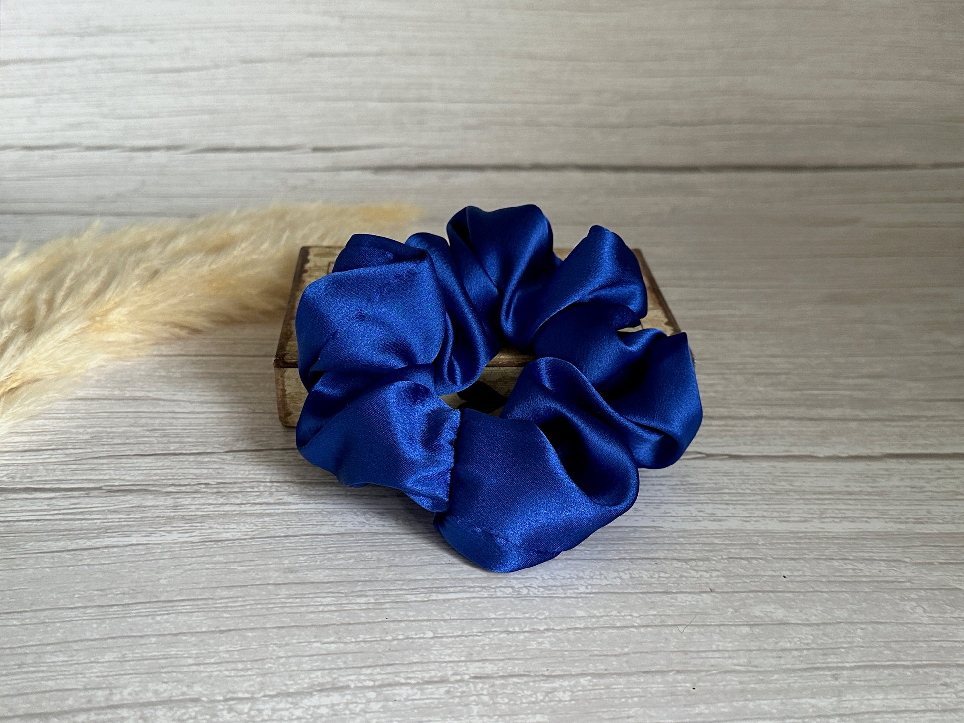 A Silk Extra Large Scrunchie in Royal Blue by Crafts by Kate rests elegantly atop a small wooden box on a light gray wooden surface, with a fluffy stem of beige pampas grass partially visible on the left side.