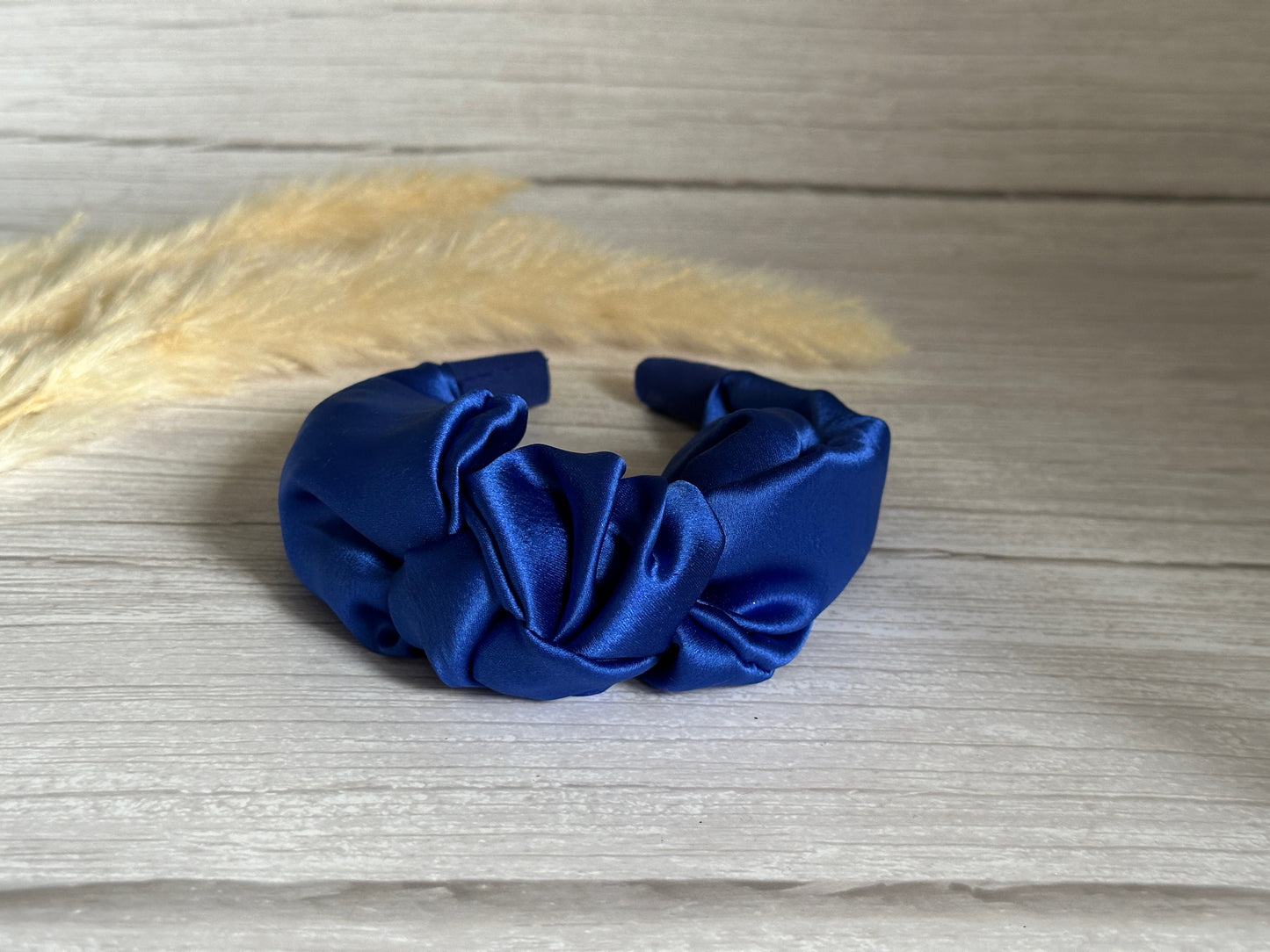 A luxurious Silk Knotted Headband in royal blue by Crafts by Kate is elegantly displayed on a wooden surface, complemented by decorative beige pampas grass in the background.