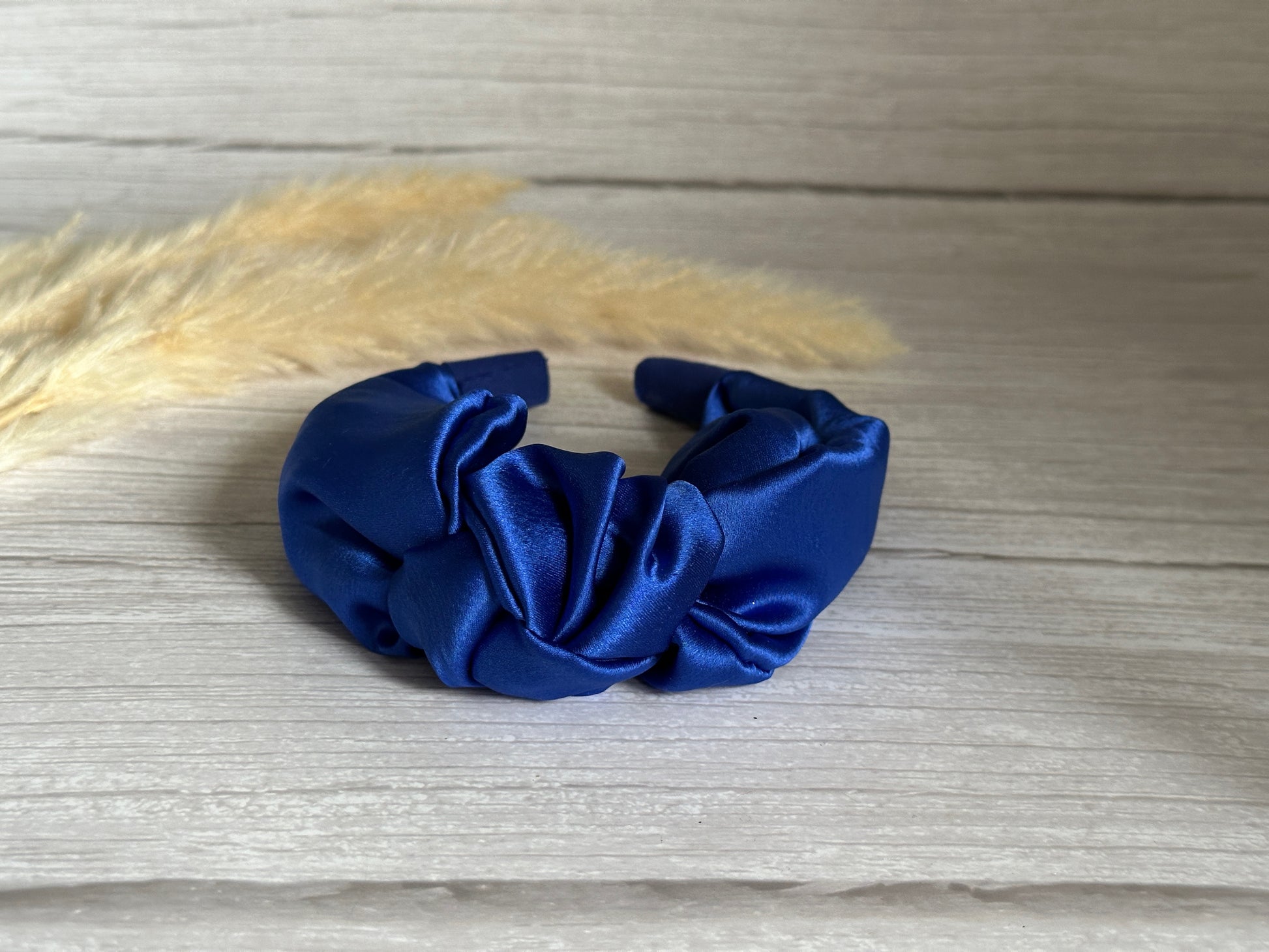 A luxurious Silk Knotted Headband in royal blue by Crafts by Kate is elegantly displayed on a wooden surface, complemented by decorative beige pampas grass in the background.