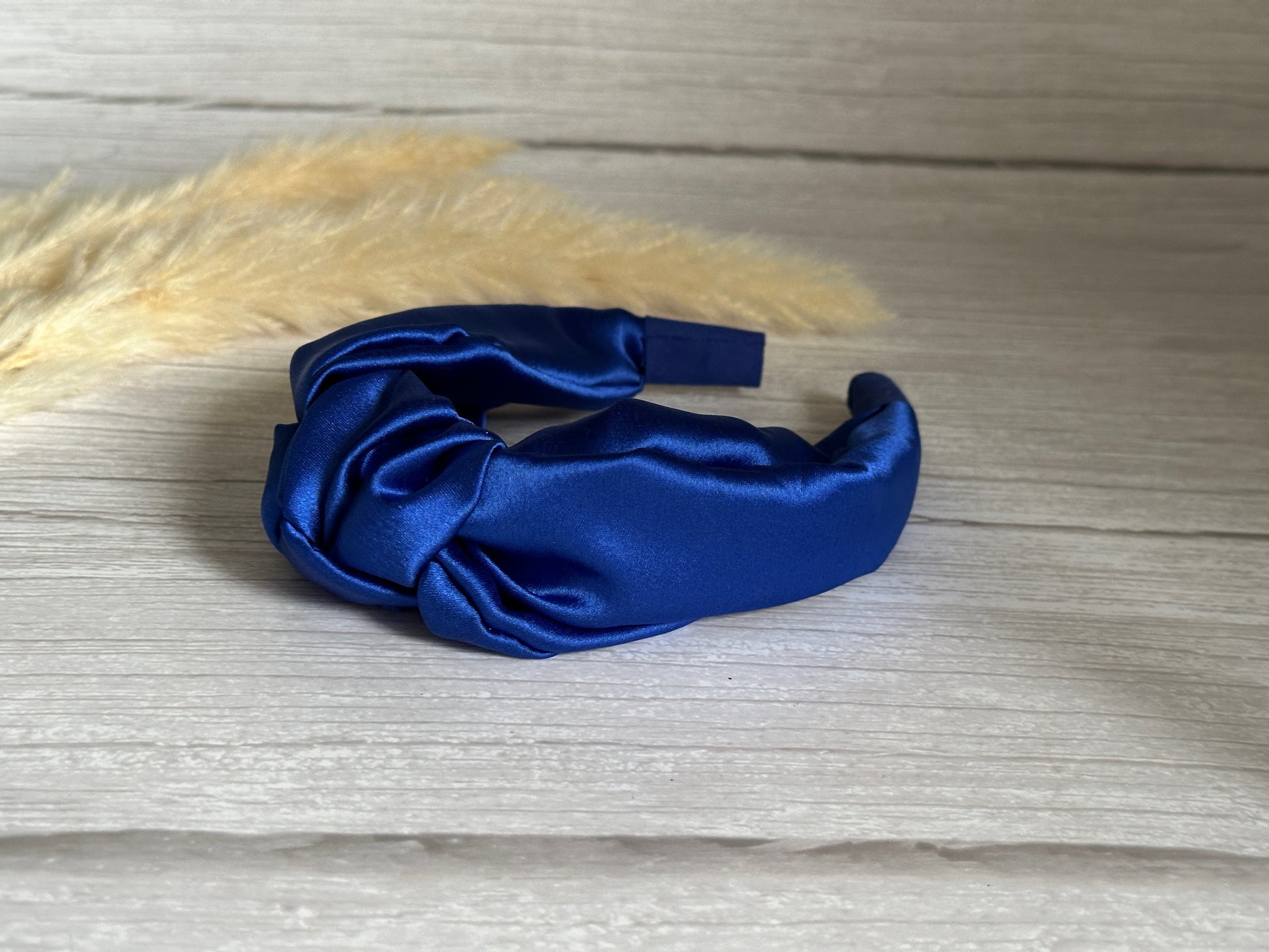 A royal blue Silk Knotted Headband from Crafts by Kate rests on a wooden surface, its large bow making a statement. To the left, beige pampas grass adds a natural touch to this luxurious composition.