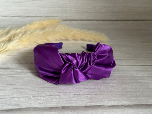 The elegant Silk Knotted Headband by Crafts by Kate, in royal purple and made from sumptuous silk with a stylish twisted knot design, is gracefully displayed on a light wooden surface. In the backdrop, two stalks of beige pampas grass contribute a natural accent to the setting.