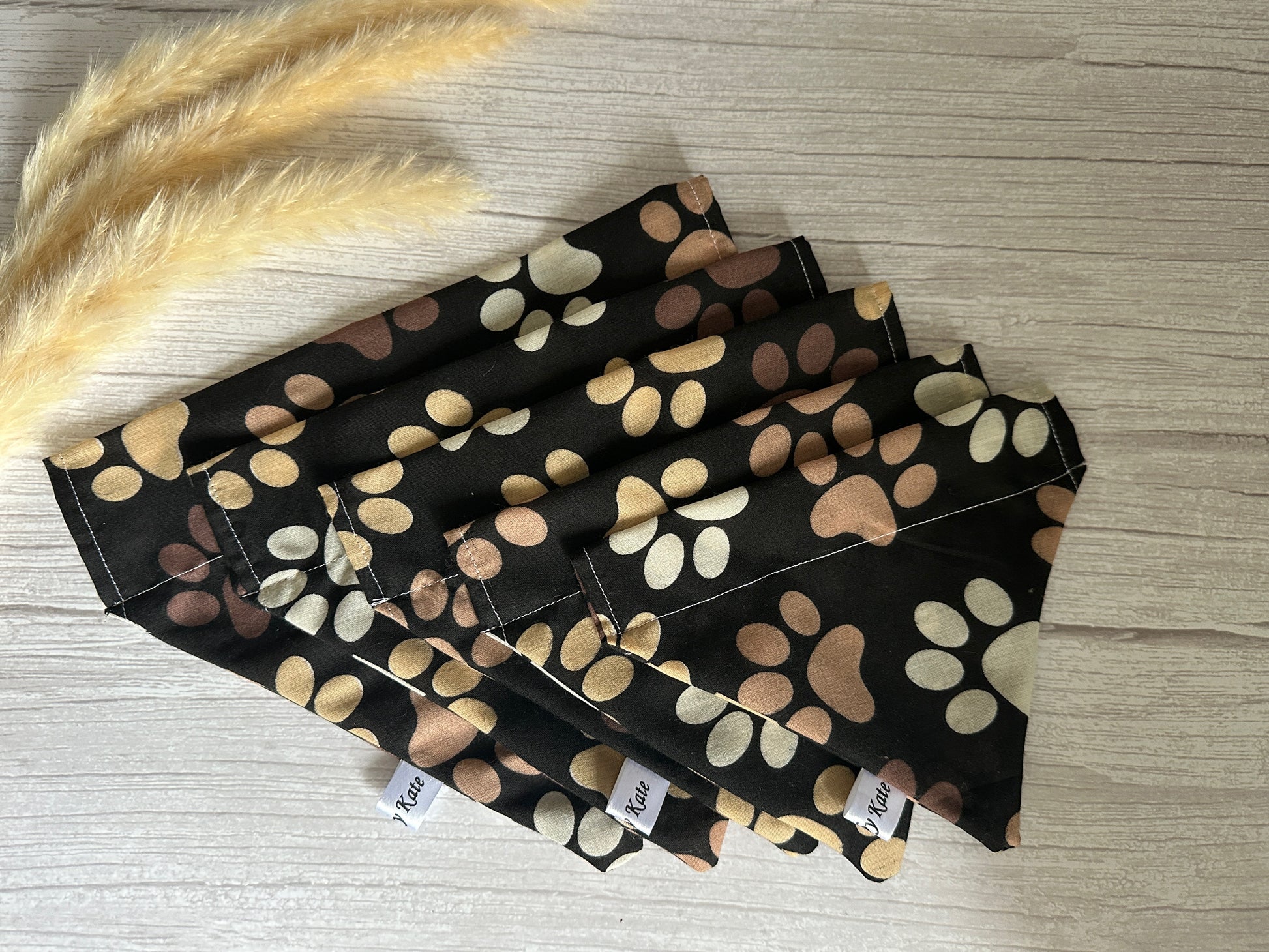 A collection of six 'Shades of Coffee' Cotton Pet Bandanas with collar attachments by Crafts by Kate is arranged on a light wooden surface. The black bandanas are adorned with a charming pattern of coffee-colored paw prints. Two small tufts of beige dried grass are positioned to the left of the bandanas.