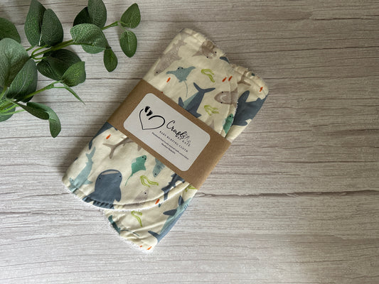 The Cotton & Terry Towelling Baby Burping Cloth - Shark Tank by Crafts by Kate, showcasing marine animal designs and crafted from ultra-soft cotton, is presented with a brown paper label and placed on a light wooden surface. Green leaves can be seen to the left.