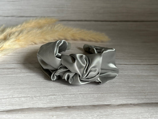 The Silk Knotted Headband - Metallic Silver by Crafts by Kate is exquisitely crafted from ruffled, metallic silver satin. It rests elegantly on a wooden surface, accented by soft beige pampas grass in the background. Its smooth, shiny texture forms graceful curves and folds.