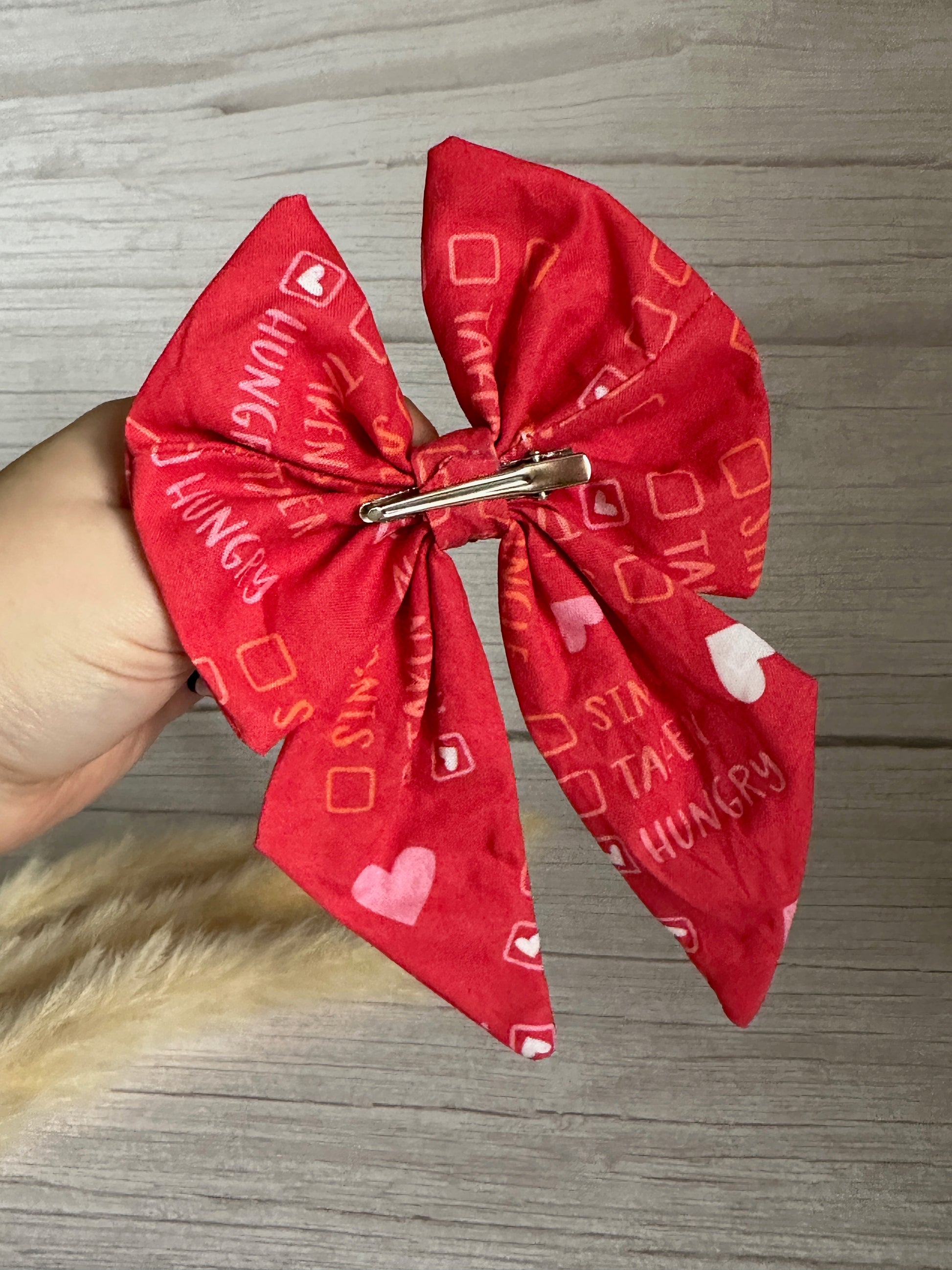 A hand holding a Cotton Sailor Bow Clip - Single/Taken/Hungry from Crafts by Kate, a handcrafted luxury red fabric bow adorned with printed hearts, squares, and phrases like "HUNGRY" and "HANGRY." The bow features a metal crocodile clip on the back. The background is a light wood surface.