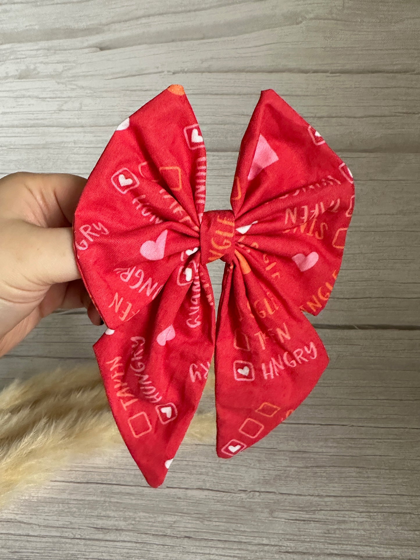 A hand is holding a large Cotton Sailor Bow Clip - Single/Taken/Hungry, handcrafted by Crafts by Kate, made of red fabric with text and small symbols printed on it, including words like "STAY HUNGRY." The background consists of light-colored wooden planks, with a furry light brown object partially visible at the bottom edge.