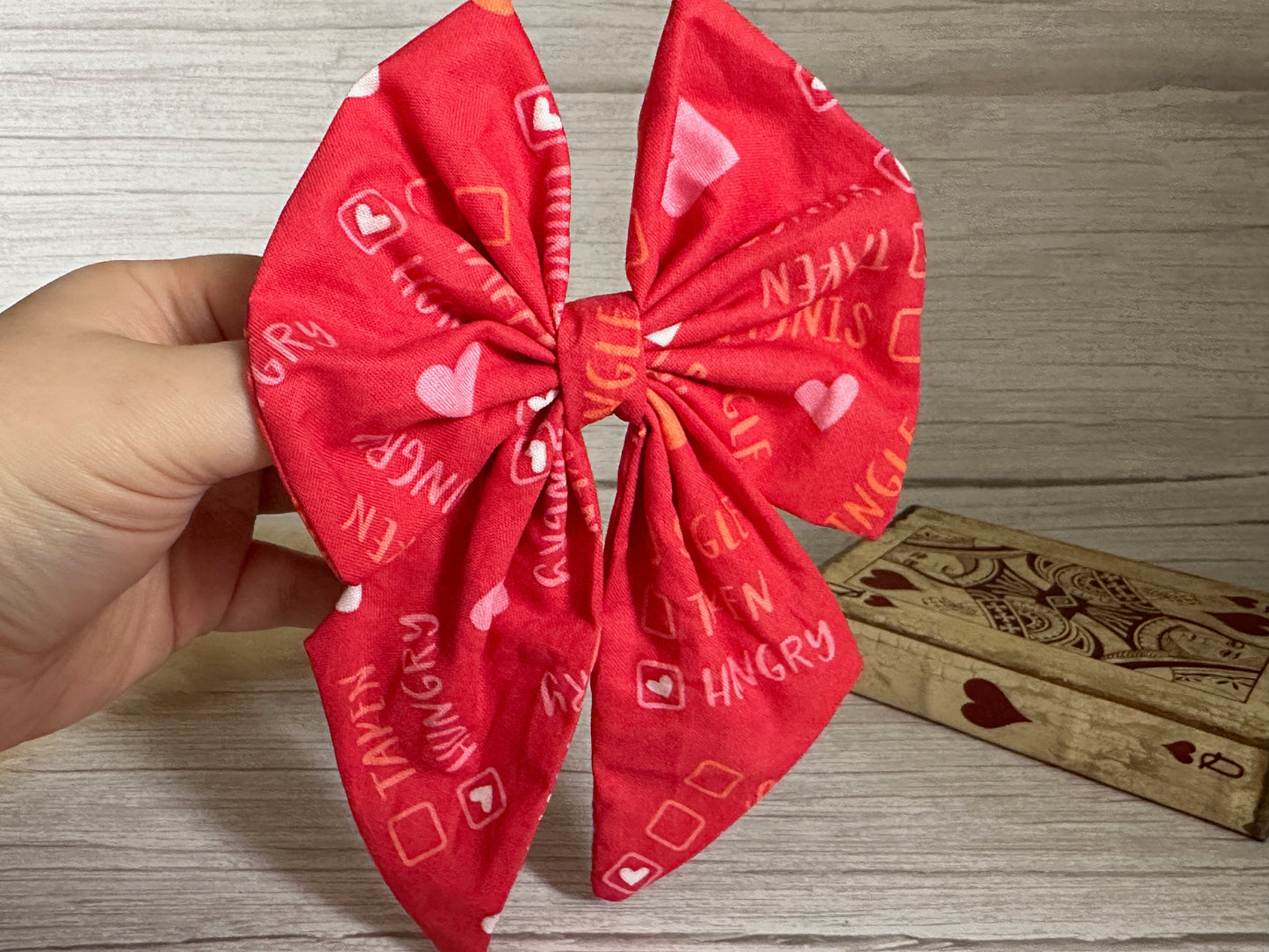 A hand is holding a large, red Cotton Sailor Bow Clip from Crafts by Kate. This handcrafted luxury bow showcases intricate heart and text patterns, including the word "HUNGRY." Behind it, there's a decorative wooden box adorned with intricate designs and a heart symbol, all set against a light wood surface.