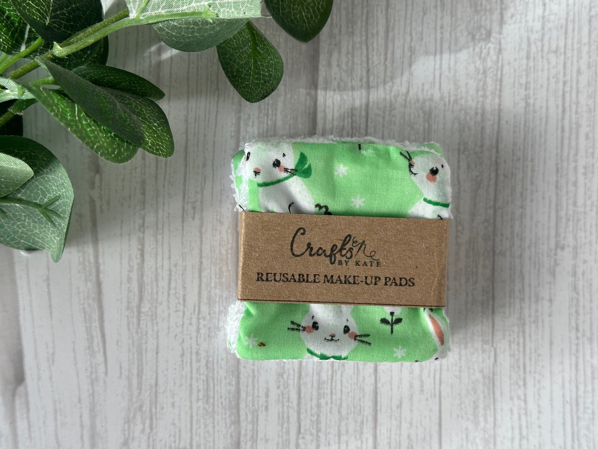 A set of Cotton Reusable Make-up Remover Pads - Spring Rabbits wrapped in brown kraft paper is adorned with "Crafts by Kate" branding. Handcrafted with a charming design featuring rabbits and animals on a green background, these environmentally friendly pads are placed on a light wooden surface near a sprig of green leaves.