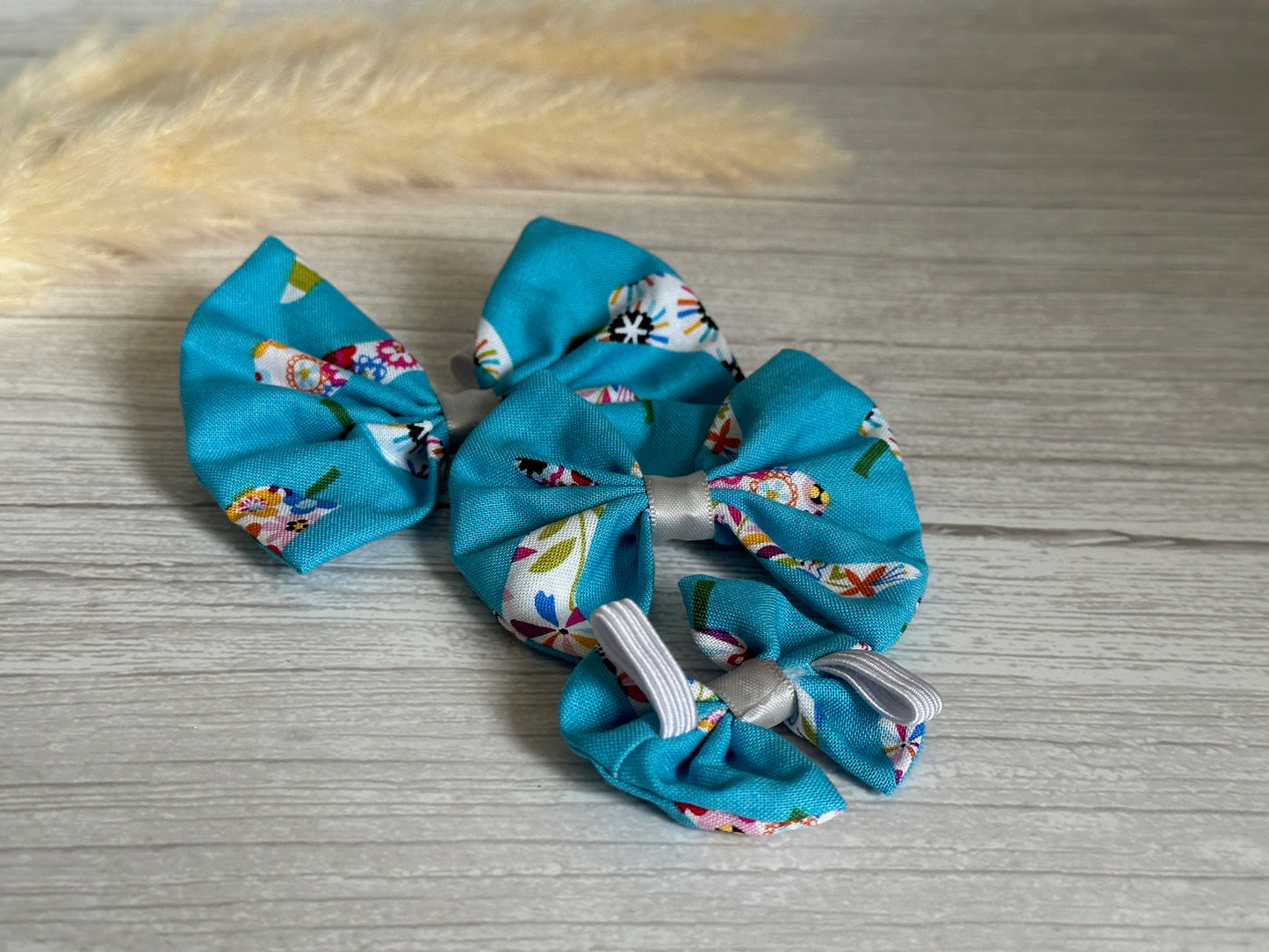 Three blue hair bows with colorful patterns, made from Summer Chilli Print Fabric by Crafts by Kate, are arranged on a gray wooden surface. In the background, a fluffy beige pampas grass decoration adds a touch of elegance. The handcrafted Cotton Pet Bows with collar attachments feature an elastic fastening for easy use.