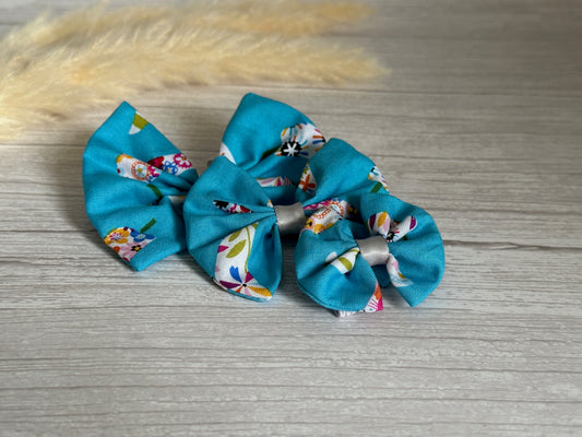 Two Cotton Pet Bows with collar attachments from the Summer Chilli collection by Crafts by Kate, featuring a blue background adorned with colorful floral patterns, lie on a light wooden surface. A dried wheat stalk is positioned in the background.