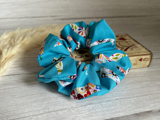 A Cotton Extra Large Scrunchie - Summer Chilli from Crafts by Kate, featuring a vibrant blue base with a floral pattern, is elegantly placed on a light wooden surface. Designed for summer, this cotton accessory pairs beautifully with a small decorative box adorned with intricate patterns subtly visible in the background.