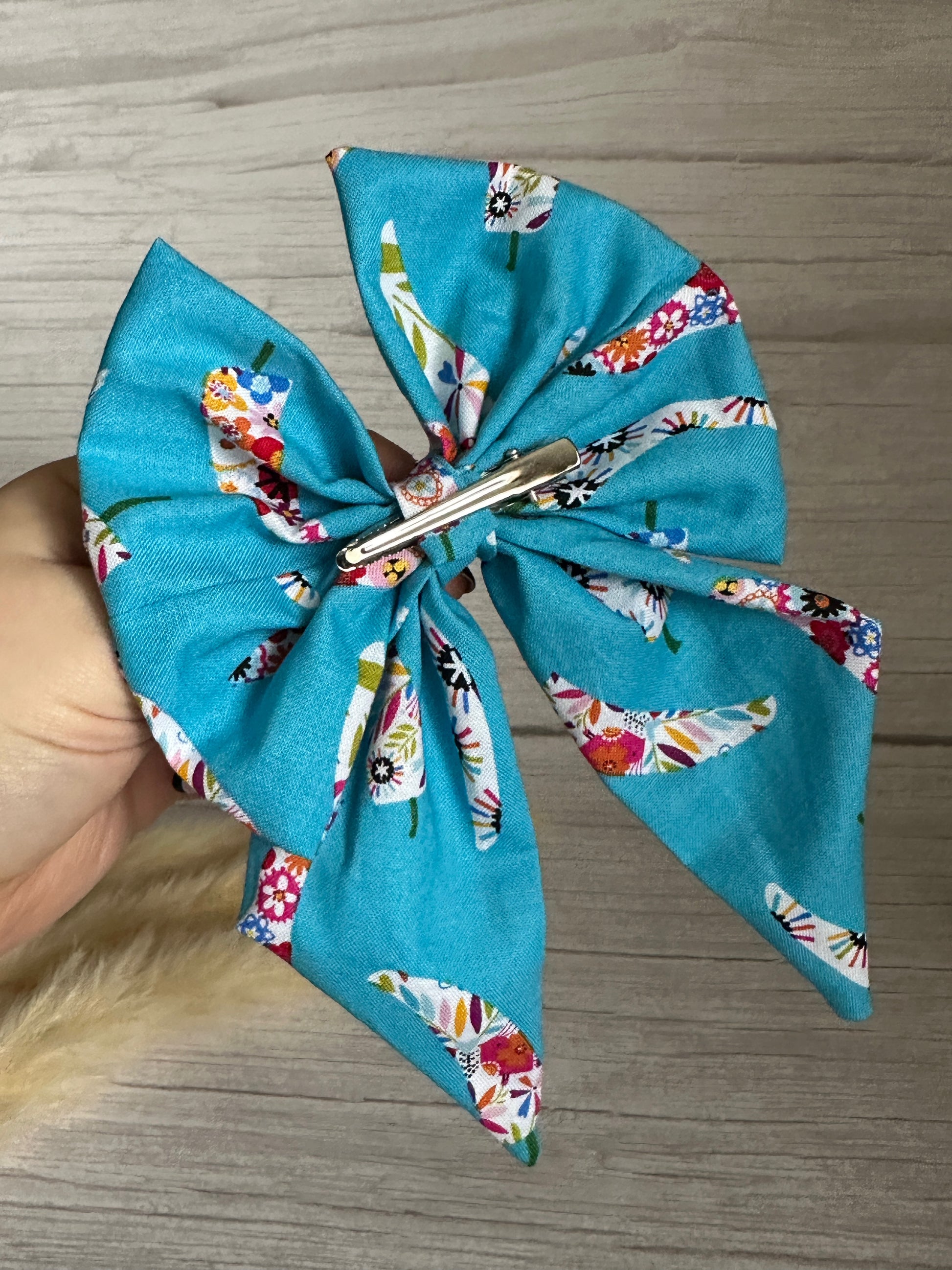 A large, handcrafted luxury Cotton Sailor Bow Clip - Summer Chilli from Crafts by Kate, made from blue cotton summer fabric featuring a colorful bird and floral pattern. The bow is attached to a silver metal clip and is shown held in hand against a light wooden surface background.