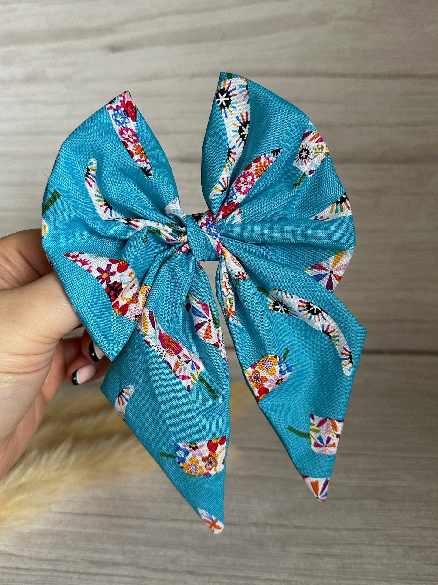 A hand holding a large, handcrafted luxury Cotton Sailor Bow Clip - Summer Chilli by Crafts by Kate, adorned with colorful floral patterns and llama designs against a rustic wooden surface background.