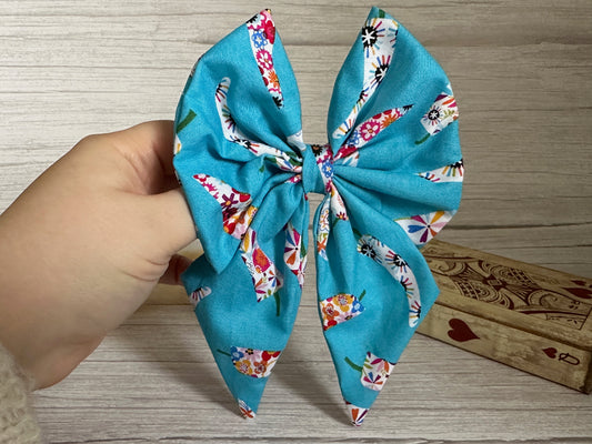 A hand is holding a large, colorful Crafts by Kate Cotton Sailor Bow Clip - Summer Chilli, made of blue fabric adorned with a pattern of vibrant flowers and shapes. In the background, two closed wooden books with red designs are partially visible on a light wooden surface.