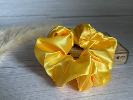 The Silk Extra Large Scrunchie in Sunshine Yellow from Crafts by Kate is elegantly displayed on a small wooden box against a light wooden backdrop. On the left, soft beige pampas grass introduces an element of texture and contrast.