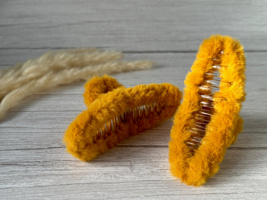 Two Fluffy Hair Claw Clips - Sunshine Yellow by Crafts by Kate are displayed on a wooden surface with dry pampas grass blurred in the background. The handcrafted clips have a soft, textured exterior, and their metal springs are visible.