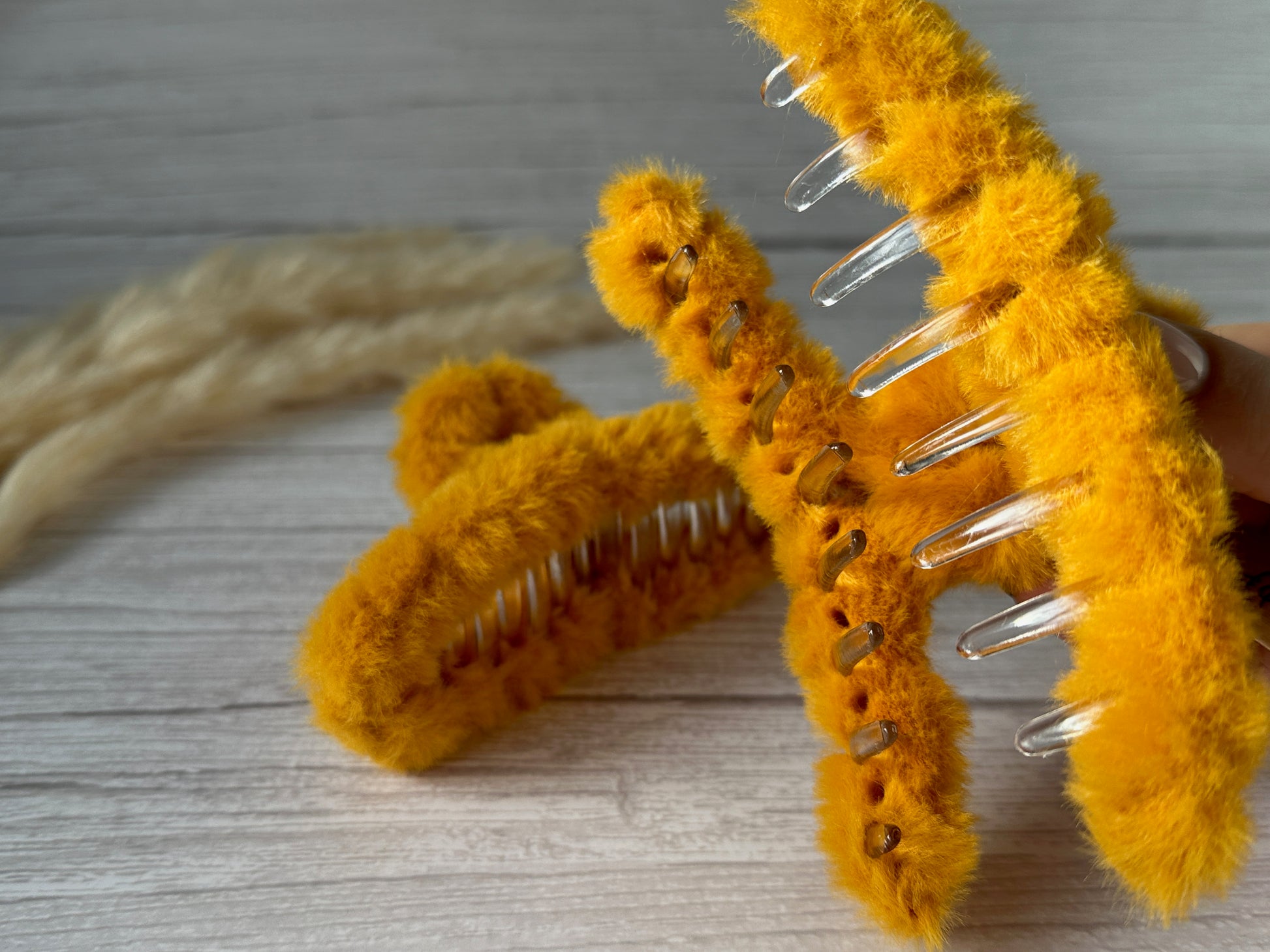 Close-up image of two Crafts by Kate "Fluffy Hair Claw Clip - Sunshine Yellow" with bright yellow fabric. The handcrafted clips have visible metal teeth, with one clip partially open and the other lying on a light wooden surface. A soft beige object is blurred in the background.