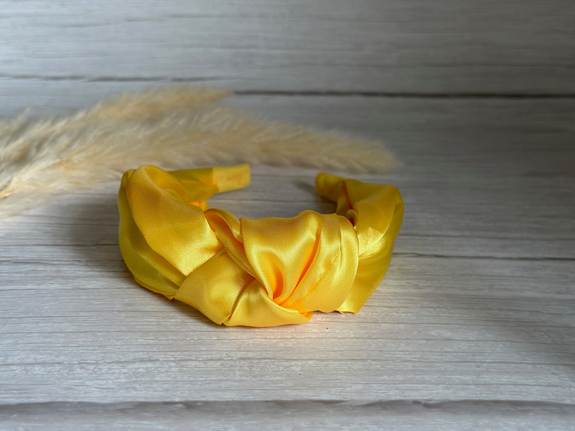 A luxurious Silk Knotted Headband in Sunshine Yellow by Crafts by Kate, featuring a twisted design, rests on a light wooden surface with soft beige pampas grass beautifully framing the background.