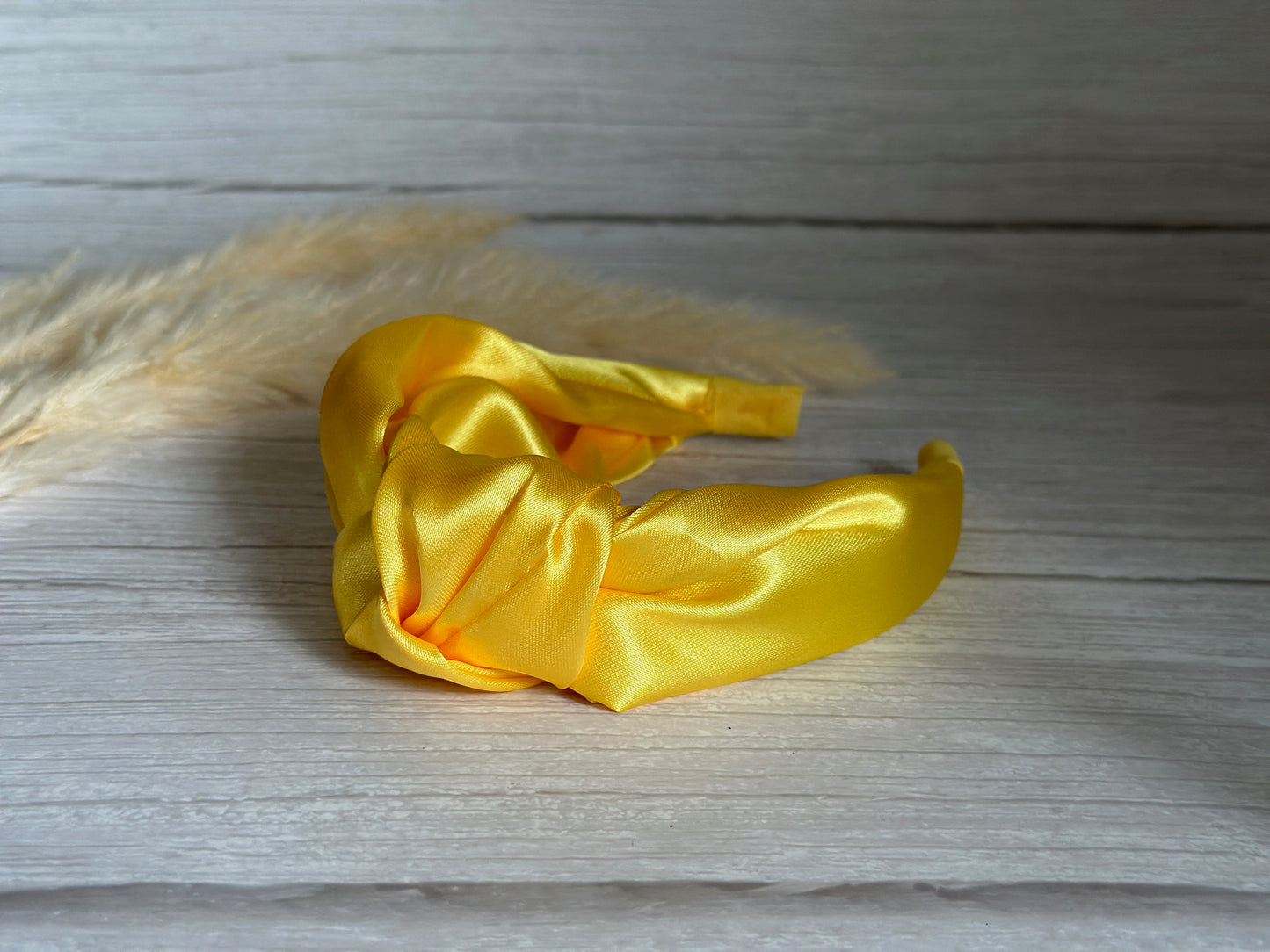 A vibrant Silk Knotted Headband in Sunshine Yellow by Crafts by Kate features a twisted knot design, resting on a light wooden surface with soft, wispy pampas grass elegantly framing the background.