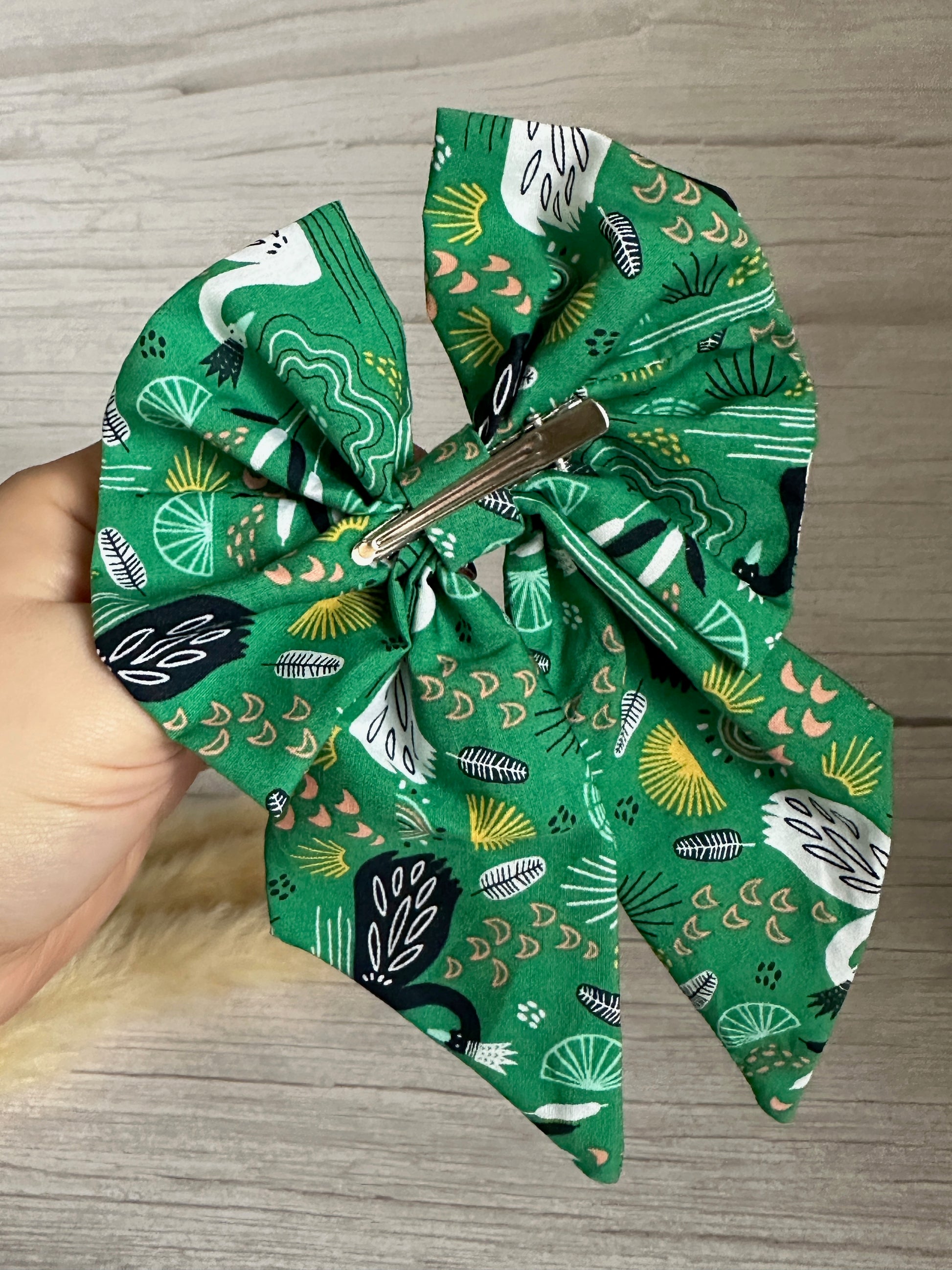 A person holds a handcrafted luxury statement piece, the Cotton Sailor Bow Clip - Swan Lake from Crafts by Kate, adorned with a nature-inspired pattern featuring leaves, birds, and other abstract designs on green fabric. The bow includes a silver metal clip on the back for attachment. It rests against a wood-textured surface background.