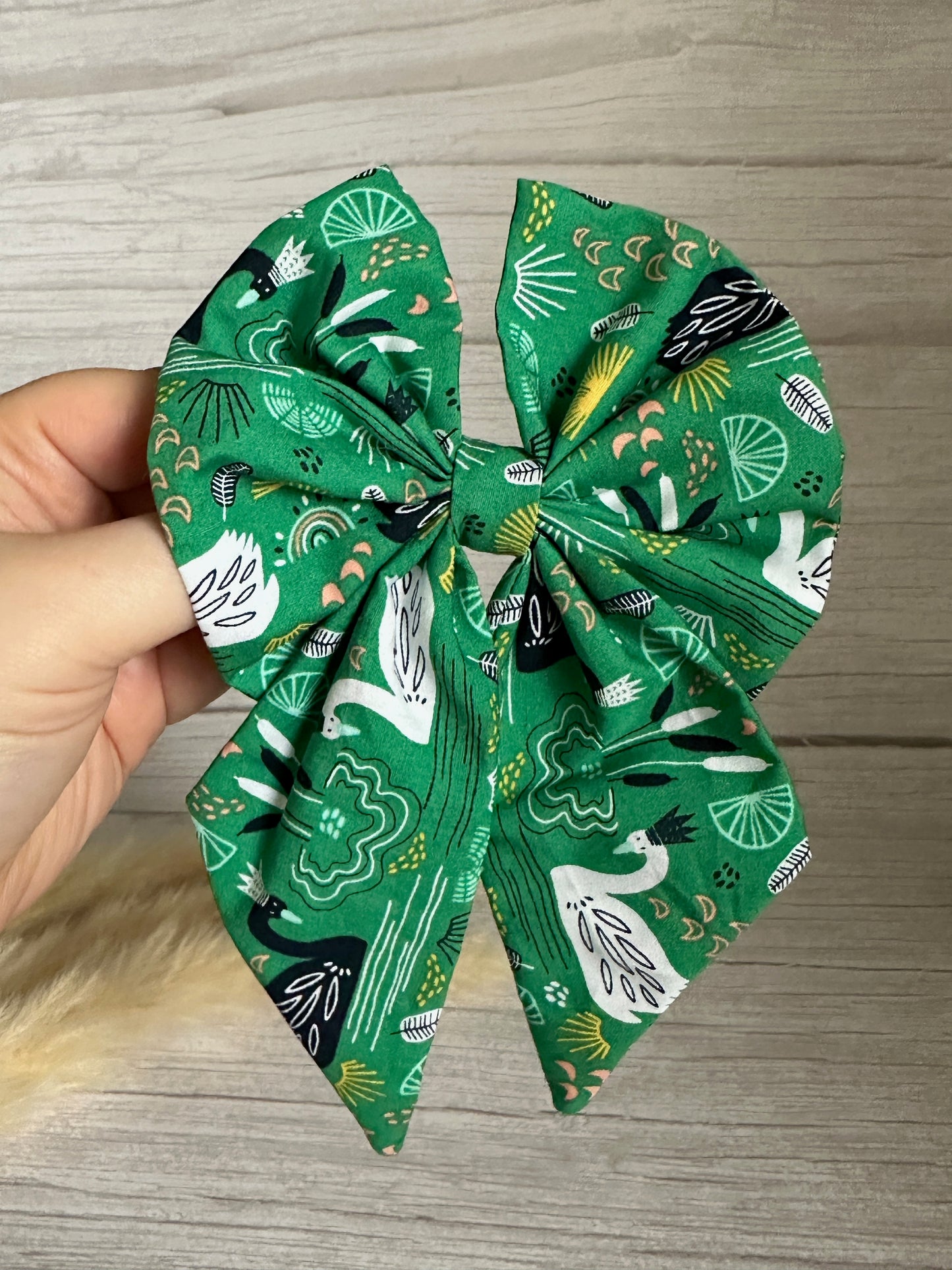 A hand holds a green cotton Sailor Bow Clip from Crafts by Kate, decorated with white birds and green leaf patterns. This Cotton Sailor Bow Clip - Swan Lake is a handcrafted luxury statement featuring two prominent loops and pointed ends, set against a light wooden background.