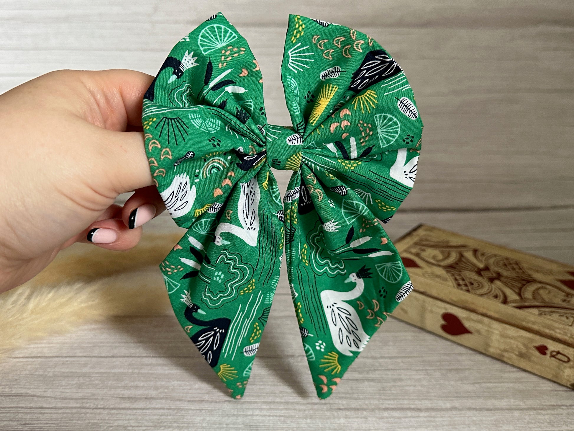 A hand is holding a green fabric Cotton Sailor Bow Clip - Swan Lake from Crafts by Kate, featuring a festive pattern with peacocks, fans, and leaves. In the background, there is a light wooden surface and a decorative wooden box adorned with a heart suit symbol. The person’s nails are painted in black and white polish.