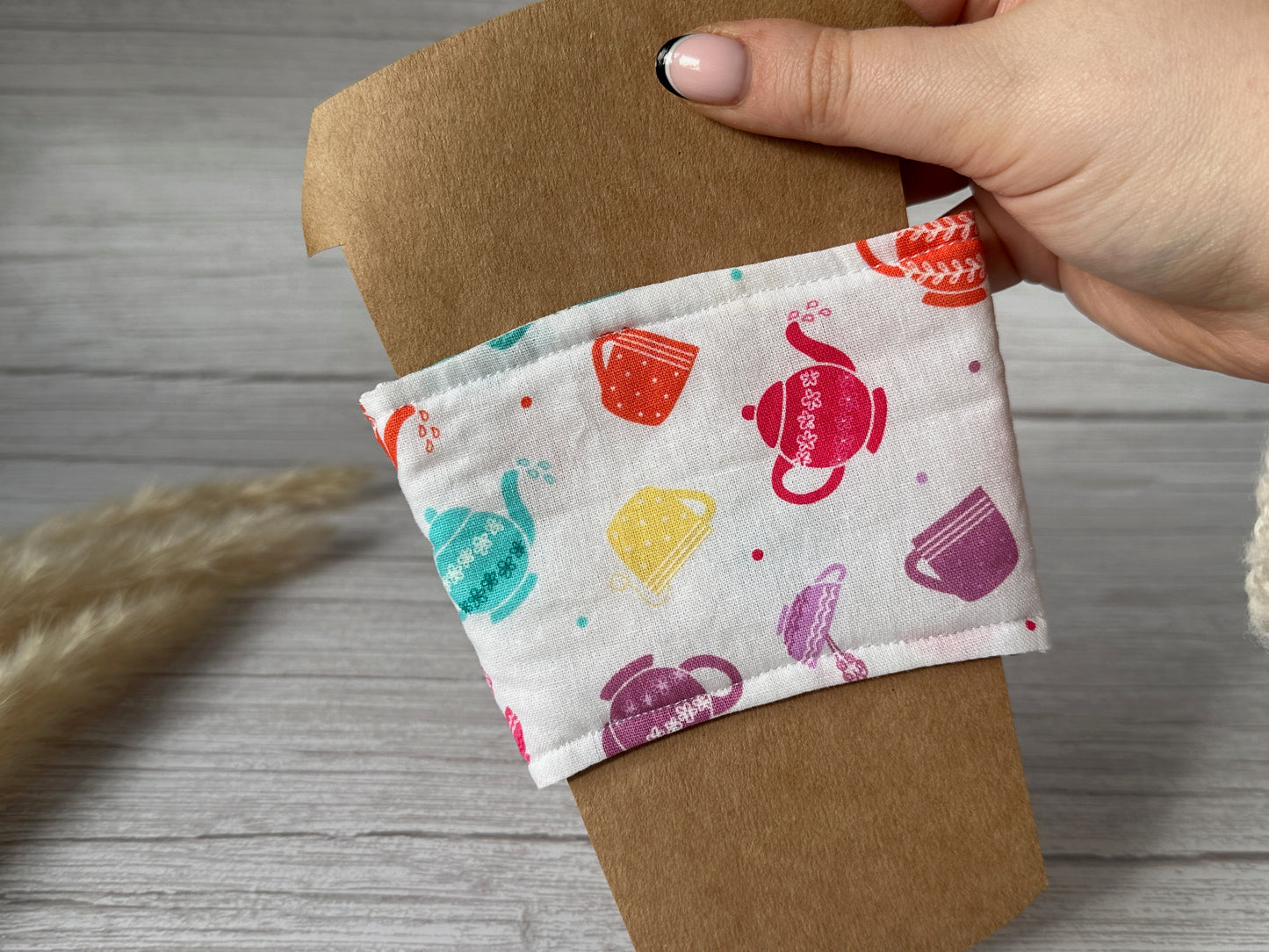 A hand with a light-colored manicure holds an environmentally friendly brown paper coffee cup adorned with the "Cotton Reusable Cup Cozy - Tea Lover" from Crafts by Kate. The handcrafted fabric sleeve is white and features colorful illustrations of teapots in red, blue, yellow, pink, and purple. The background includes a light wooden surface with some dried grass on the left side.