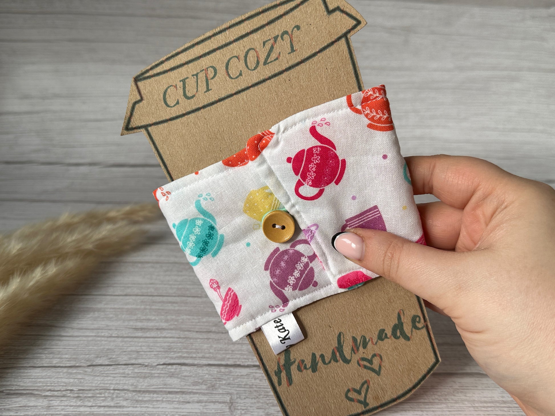 Close-up of a hand holding the Crafts by Kate Cotton Reusable Cup Cozy - Tea Lover, featuring colorful teapot designs and a wooden button on the front. The cozy is displayed against a cardboard cutout shaped like a take-out coffee cup, labeled "Cup Cozy." The background is a light wood surface.