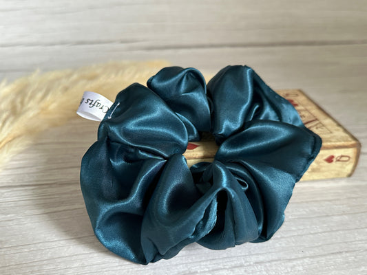 A luxurious teal blue satin scrunchie from Crafts by Kate is elegantly placed on a wooden surface, while an old, closed book adorned with playing card symbols can be seen nearby.