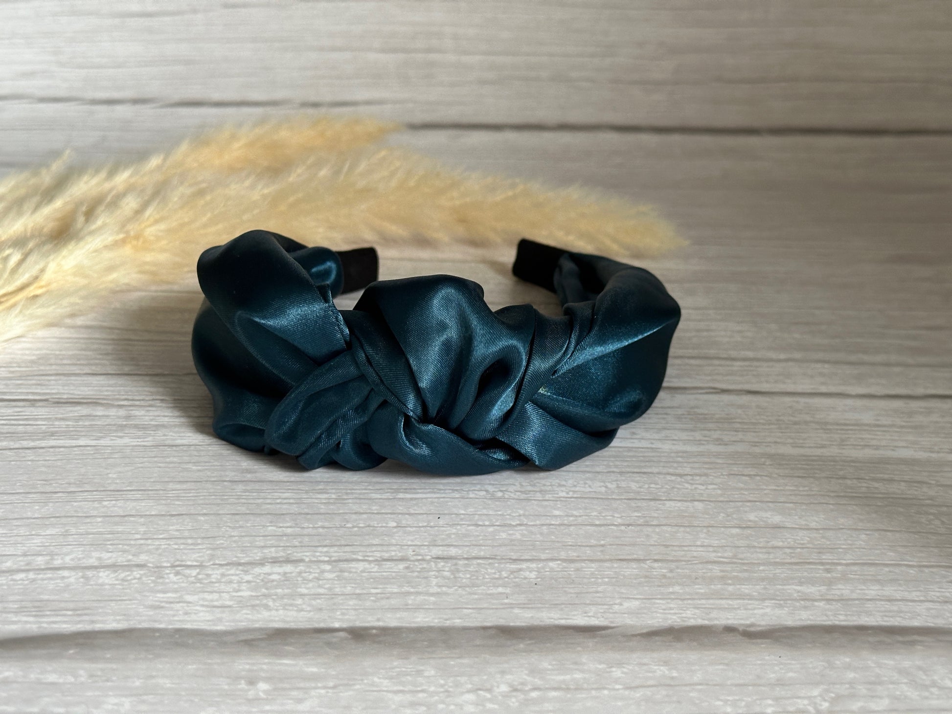 A silk knotted headband in an elegant teal blue by Crafts by Kate is placed on a light wooden surface, beautifully complemented by dried beige grass stems.